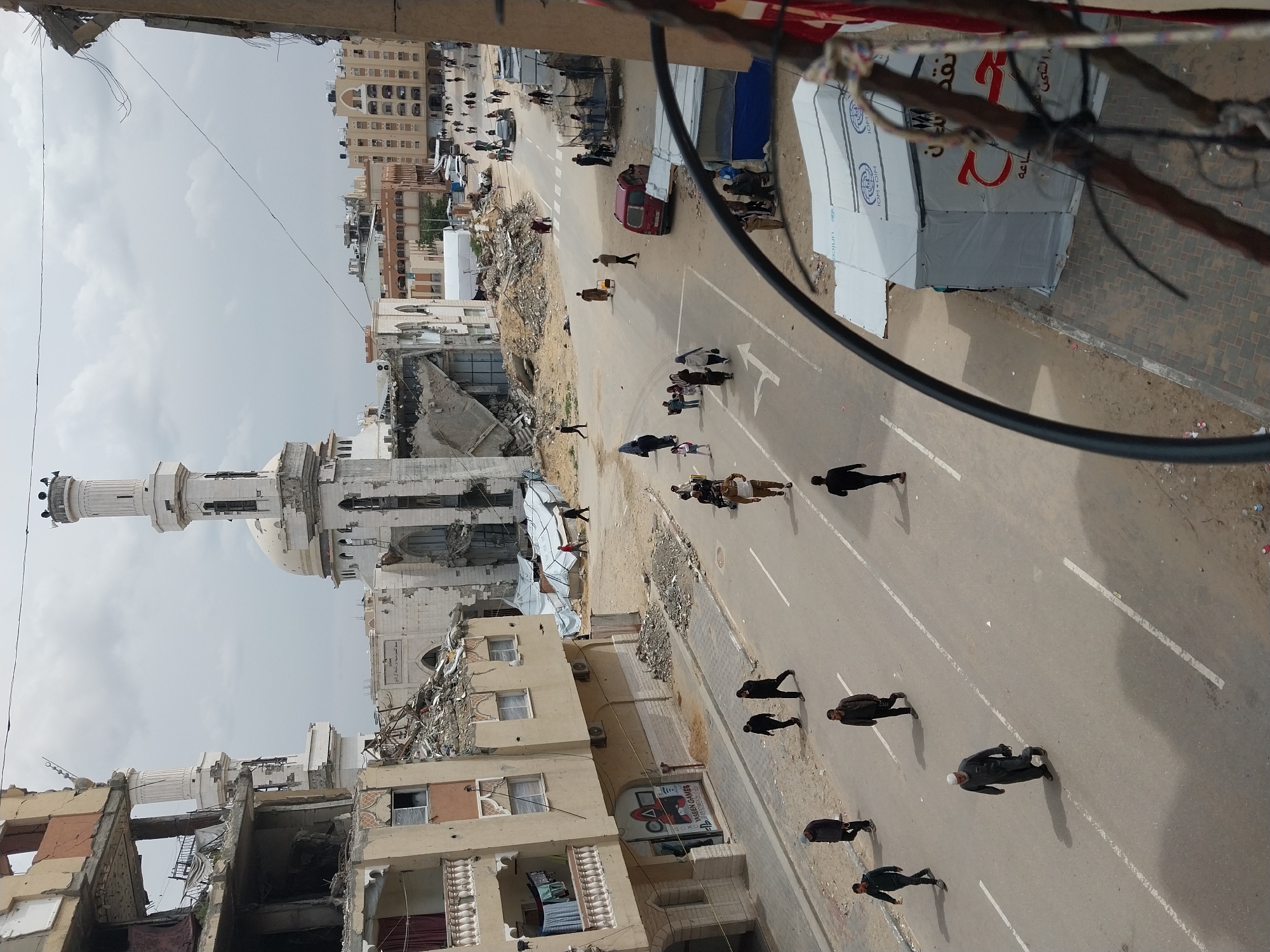 The image depicts an elevated view of a main street in Khan Yunis, Palestinian Territory, characterized by significant urban damage. An overcast sky looms above.
In the center-background, a prominent mosque with a large white dome and a tall, slender minaret topped by a multi-tiered crown and loudspeakers stands damaged, with portions of its facade missing and rubble accumulated around its base. Adjacent structures near the mosque also display extensive damage.
To the left, a multi-story building shows severe structural compromise, with upper levels partially collapsed, exposing rebar and interior elements. A lower floor features balconies, with laundry visible on one. The ground floor of this building houses a storefront with "YASEEN GAMES" and "PLAYSTATION GAMES" branding, including a graphic reminiscent of "Squid Game."
The wide, asphalt street, marked with white dashed and solid lines, as well as a left-turn arrow, dominates the midground. Piles of concrete rubble and debris are scattered along the sides of the road.
Numerous pedestrians, including men, women, and children, are walking along the street and adjacent dirt and tiled sidewalks. Their attire is typical civilian clothing; some women wear head coverings. One individual in light brown robes is observed pushing a small cart along the road.
On the right side of the street, multiple humanitarian tents are visible on the pavement. These include white tents displaying "IOM OIM UNICEF" logos and at least one blue tent. A dark red van is parked near these tents. Further down the street, a black sedan is also present.
Overhead, thin cables or wires crisscross the sky. In the immediate foreground, a thick, dark cable runs diagonally from the upper left to the lower right, partially obscuring the view of the lower right section of the scene.