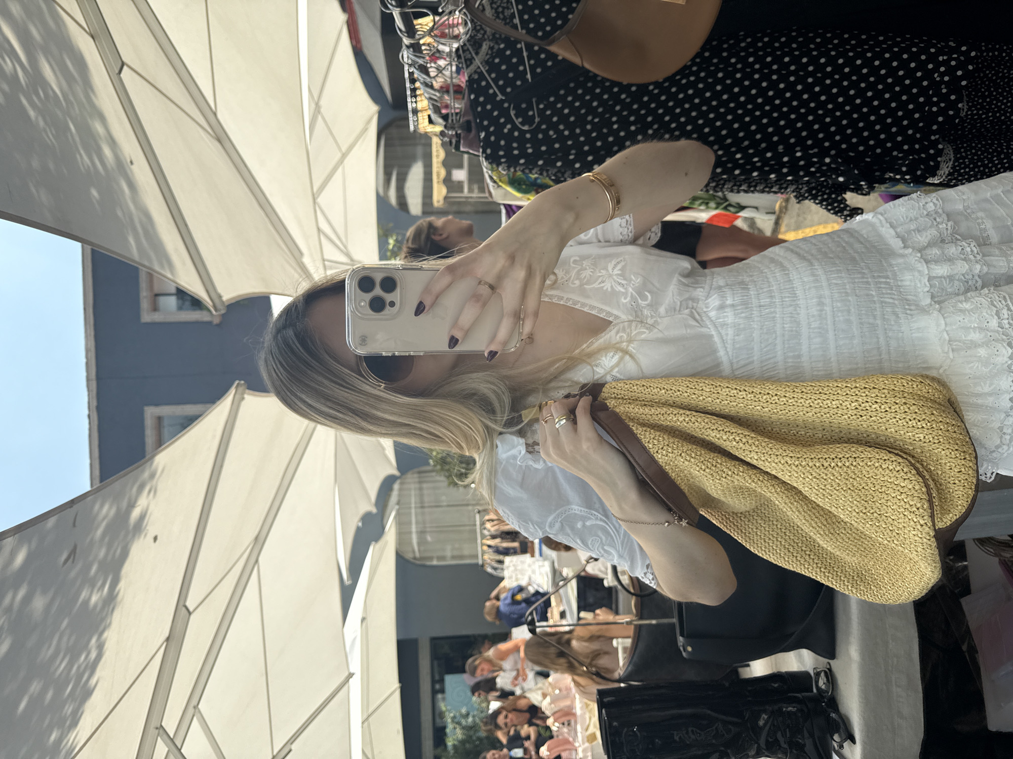 The image shows a woman taking a selfie at a market, possibly in Mexico City, Mexico. She is wearing a white dress and holding a straw bag. The background shows market stalls with various items for sale and other people browsing. White umbrellas are visible above the market stalls.