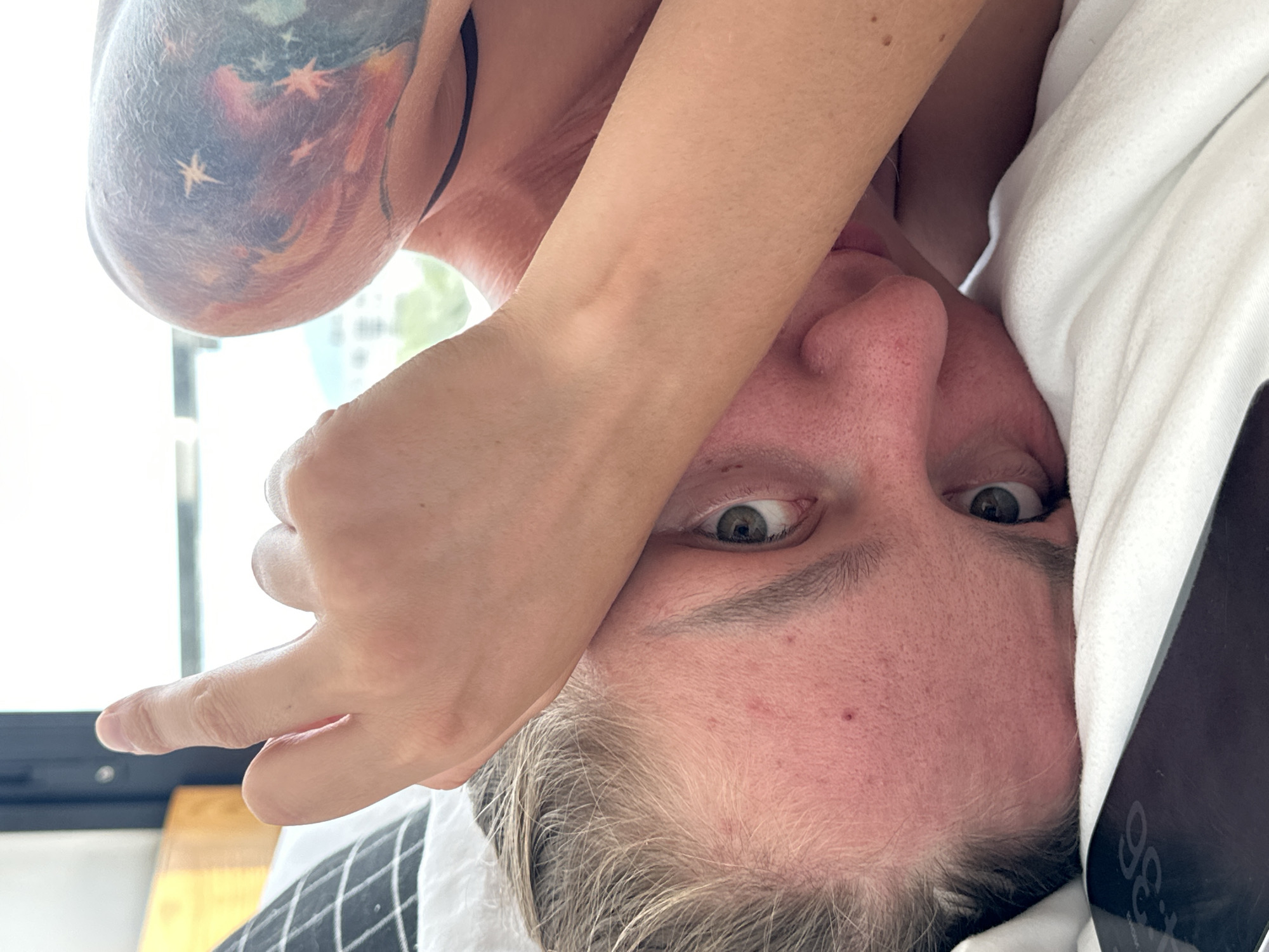 The image shows a close-up of a person lying in bed, with their face partially obscured by their arm. The person has fair skin, light-colored hair, and green eyes. They are wearing a black bra strap visible around their neck. A tattoo of a celestial scene, featuring stars and nebulae, is visible on their shoulder. A phone with the time "4:36" displayed is on the bed next to them. The background includes a window with natural light streaming in, suggesting an indoor setting. There is no visual information in the image that would allow for a specific location in the city of Đà Nẵng, Vietnam.