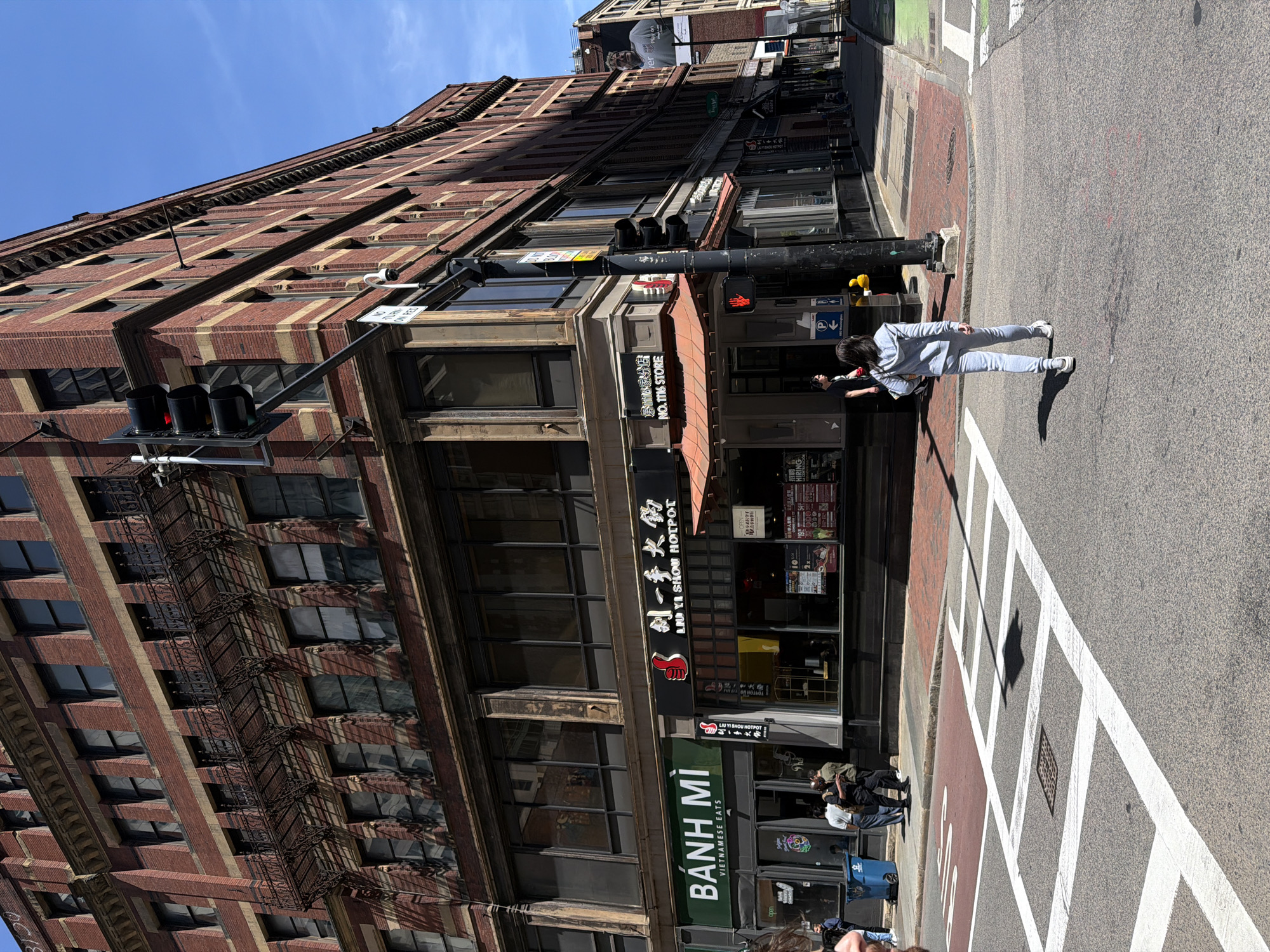 The image depicts a street corner in Boston, Massachusetts, United States. A tall, brick building dominates the left side of the frame. The building is several stories high and features a fire escape on its side.
At street level, the storefronts display signs for restaurants, including "BÁNH MÌ" (Vietnamese eats) and "LIU YI SHOU HOTPOT" (in both English and Chinese characters).
A woman is standing on the crosswalk in the street, with her arms outstretched. The street itself shows painted white crosswalk lines and a portion of the roadway surface. The sky is visible in the background, appearing blue and clear.