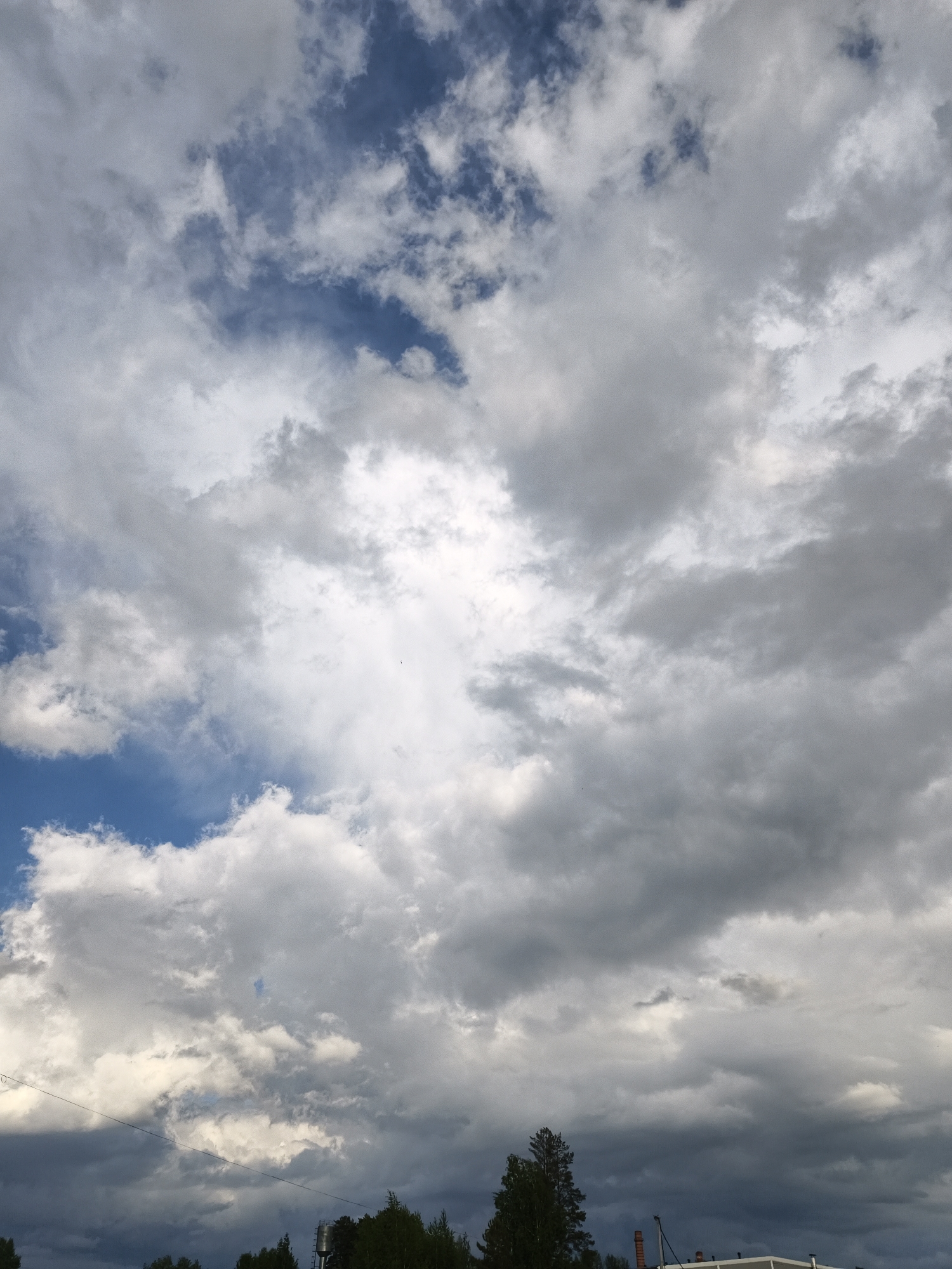The image depicts a cloudy sky with patches of blue visible. The clouds are a mix of white and grey, suggesting varying levels of thickness and moisture. At the bottom of the frame, trees and a building are partially visible. The location is Noviy, Russia. The environment suggests an outdoor setting with overcast weather conditions. No people are visible. The scene is static, capturing a moment in time.