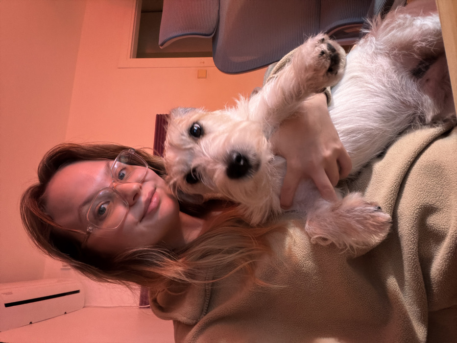 A woman with glasses smiles, holding a fluffy white dog. She wears a tan hoodie. The dog has shaggy fur and dark eyes. The background is peach-colored, with a window and a chair visible. An air conditioning unit is seen on the floor. The image is angled, creating a dynamic perspective.