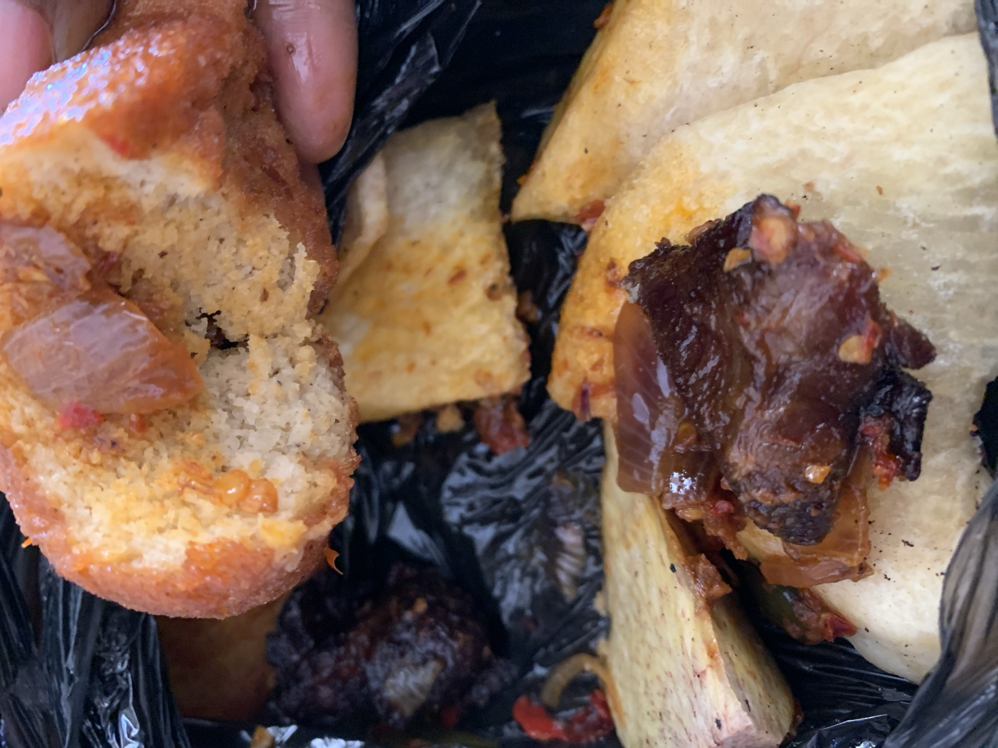 The image shows discarded food items in a black plastic bag. A piece of what appears to be fried bread, torn open, is held by a hand. Within the bag, there are pieces of what look like fried yam and dark, possibly burnt, pieces of meat or vegetables, seemingly seasoned with peppers and onions. The environment suggests a lack of formal waste disposal facilities. The image does not provide sufficient information to confirm it was taken in Kabong, Nigeria.