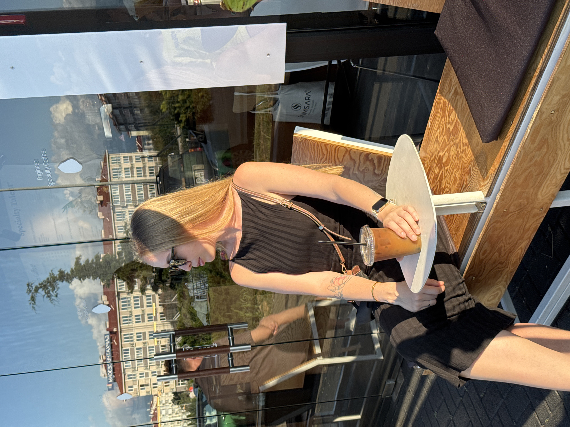 In Sochi, Russia, a blonde woman is seated at an outdoor cafe table. She is wearing a black dress and sunglasses, and is holding a cup of iced coffee. She has a tattoo on her arm and is wearing a bracelet and watch. In the reflection on the glass window behind her, buildings and trees are visible. A shopping bag with the Samsara logo is also visible in the reflection.