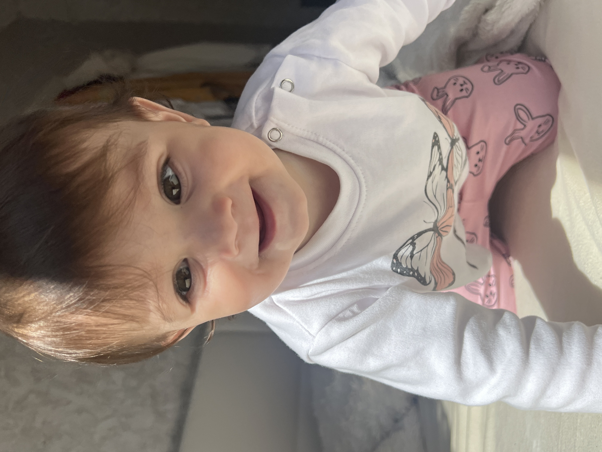 A baby girl, approximately 6 to 12 months old, is shown from a high-angle, close-up view. She has fair skin, brown hair, and brown eyes. She is wearing a white, long-sleeved shirt with a butterfly design on the front. Underneath the shirt, she is wearing pink pants with a bunny pattern. The background is blurred and indistinct, but appears to be a room with light-colored walls and some furniture. There is no indication the image was taken in Elena, Bulgaria.