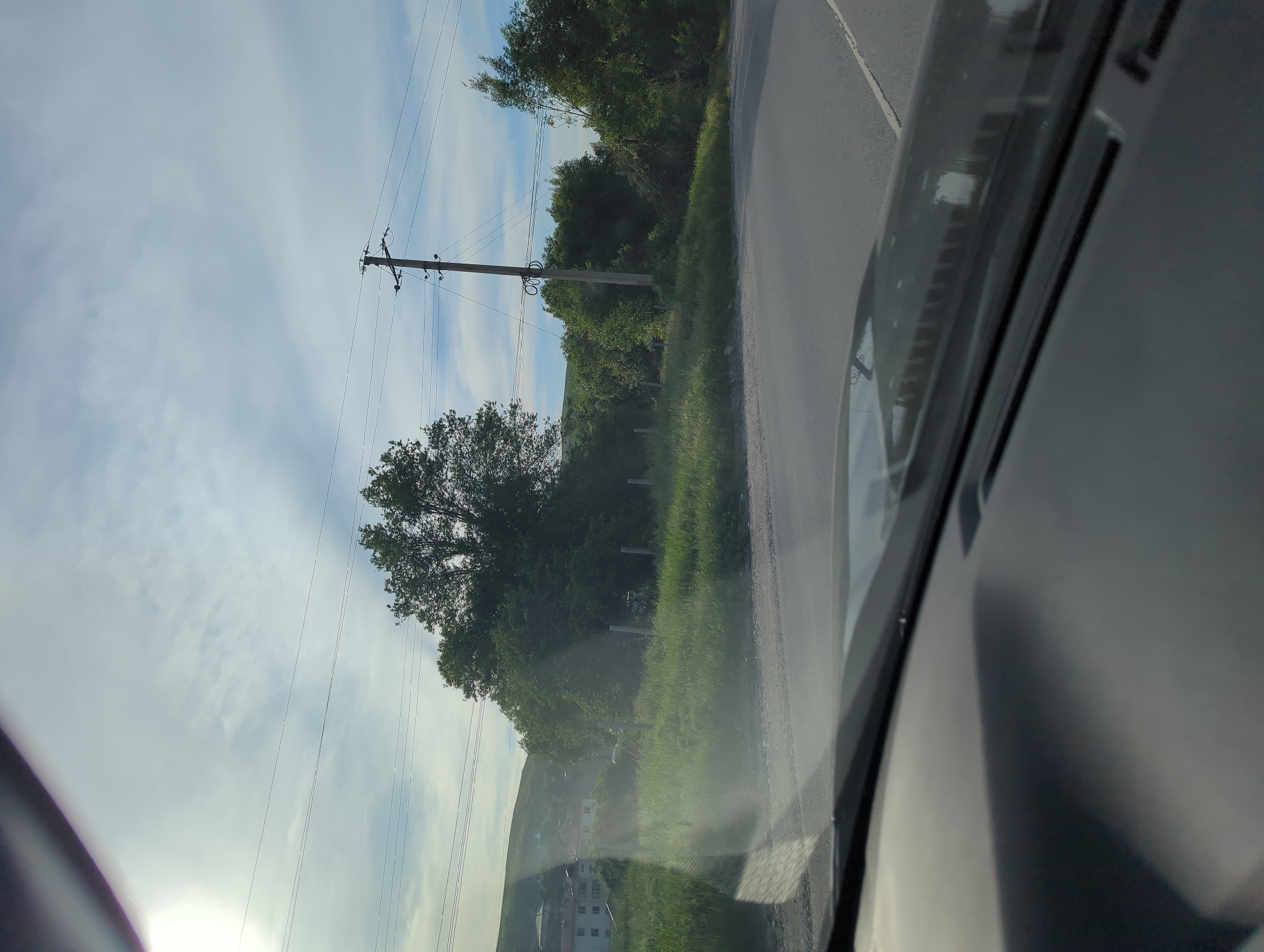 View from inside a vehicle traveling along a road in Beş-Küŋgöy, Kyrgyzstan. The road runs parallel to a grassy area with intermittent fencing and trees. Overhead power lines run alongside the road. A large tree is prominently visible on the left side of the road, with buildings visible in the background. The sky is partly cloudy. The dashboard and windshield of the vehicle are visible in the foreground.