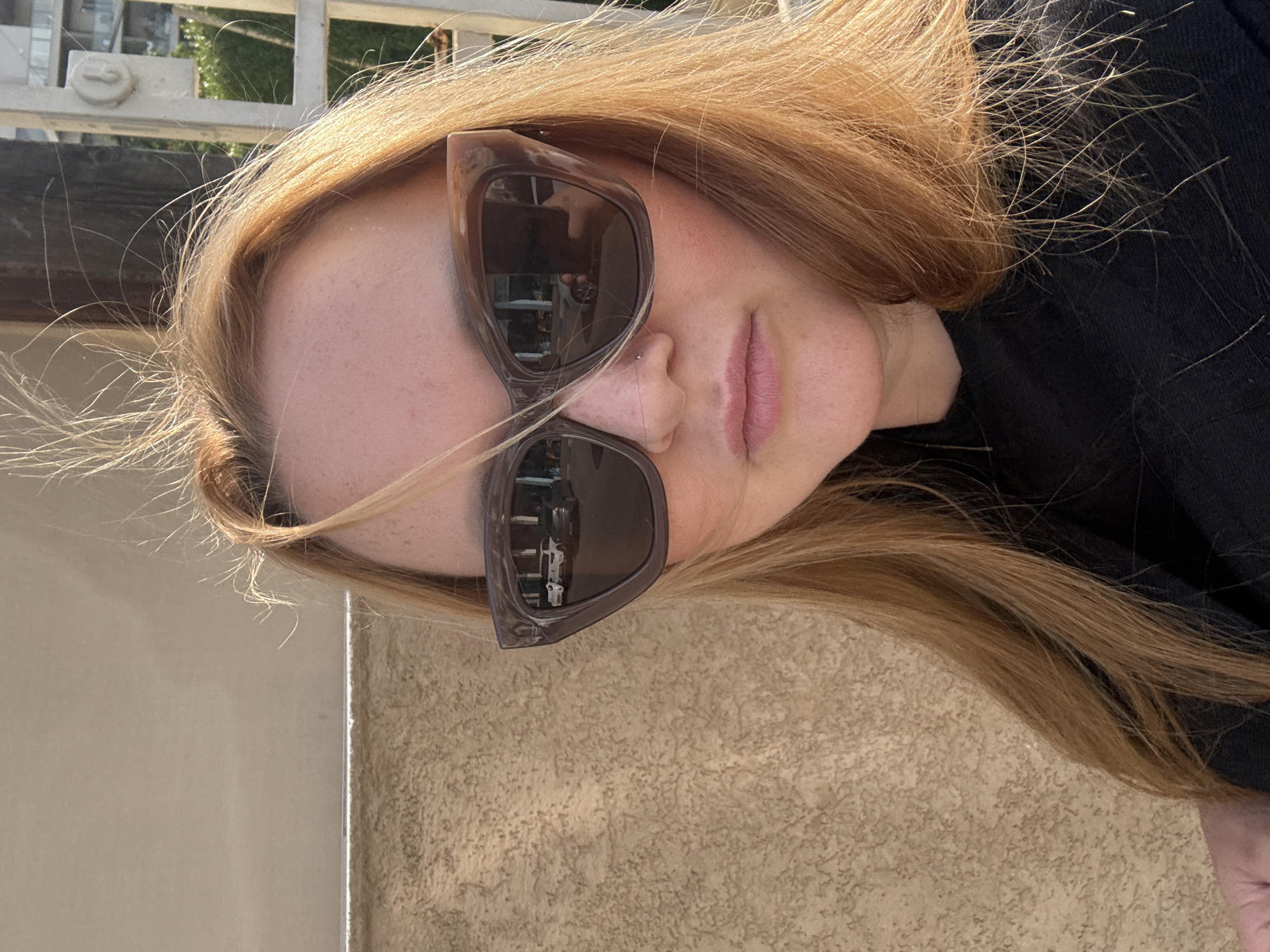 A rotated, close-up portrait shot of a person, likely a woman, with long, light brown or blonde hair in an outdoor setting in Ramat Gan, Israel. The strong, natural light suggests it is daytime, with direct sunlight highlighting the hair and skin. The person is wearing large, dark-framed cat-eye sunglasses, which reflect elements of the surroundings, possibly an interior space with shelves or a window. A small stud nose piercing is visible on her right nostril, and her lips are a soft pink. Her fair skin shows visible freckles on her forehead and cheek. She is dressed in a dark-colored top, possibly black. In the background, to her lower right, is a textured light brown wall, and to her upper left, a smoother, lighter-toned wall. A metallic railing or fence is faintly visible in the upper left corner and through her hair, suggesting an outdoor location such as a balcony or patio. The overall mood is neutral. No text is visible in the image.