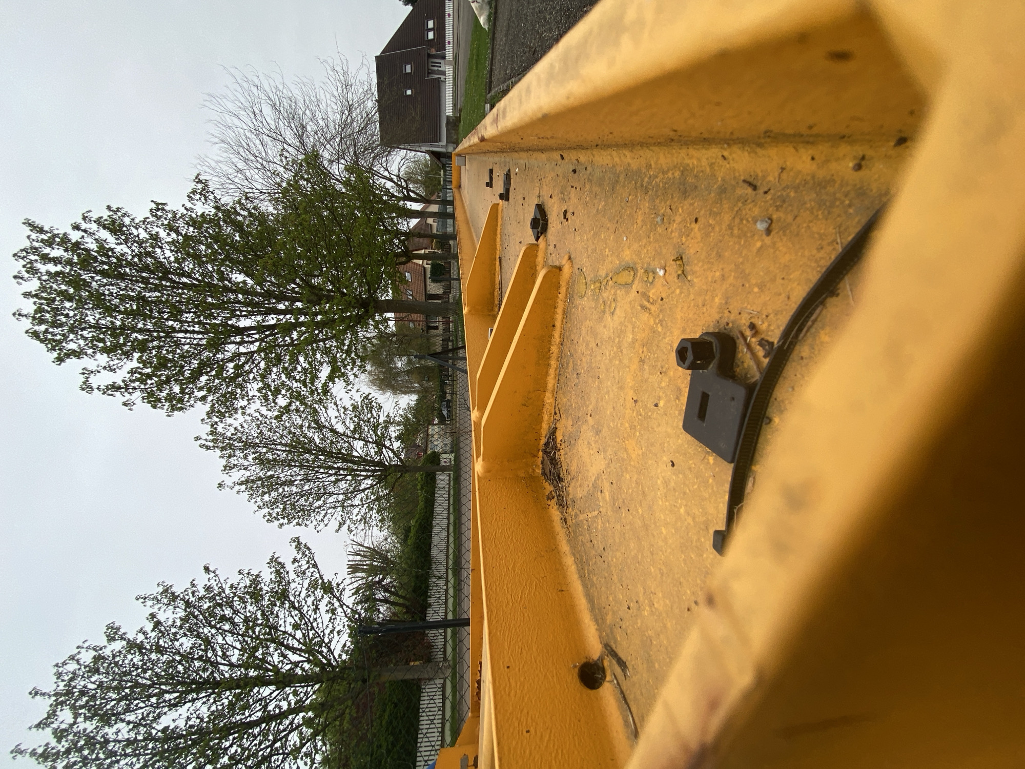 A close-up, skewed perspective of a yellow metal structure, possibly part of a vehicle or machinery, dominates the foreground. The structure shows signs of wear and tear. Through the structure, a view of a residential area in Truchtersheim, France, can be seen. Trees with varying degrees of foliage are interspersed among the houses, some of which have red-tiled roofs. A fence lines part of the scene. The sky is overcast, suggesting a cloudy or rainy day.