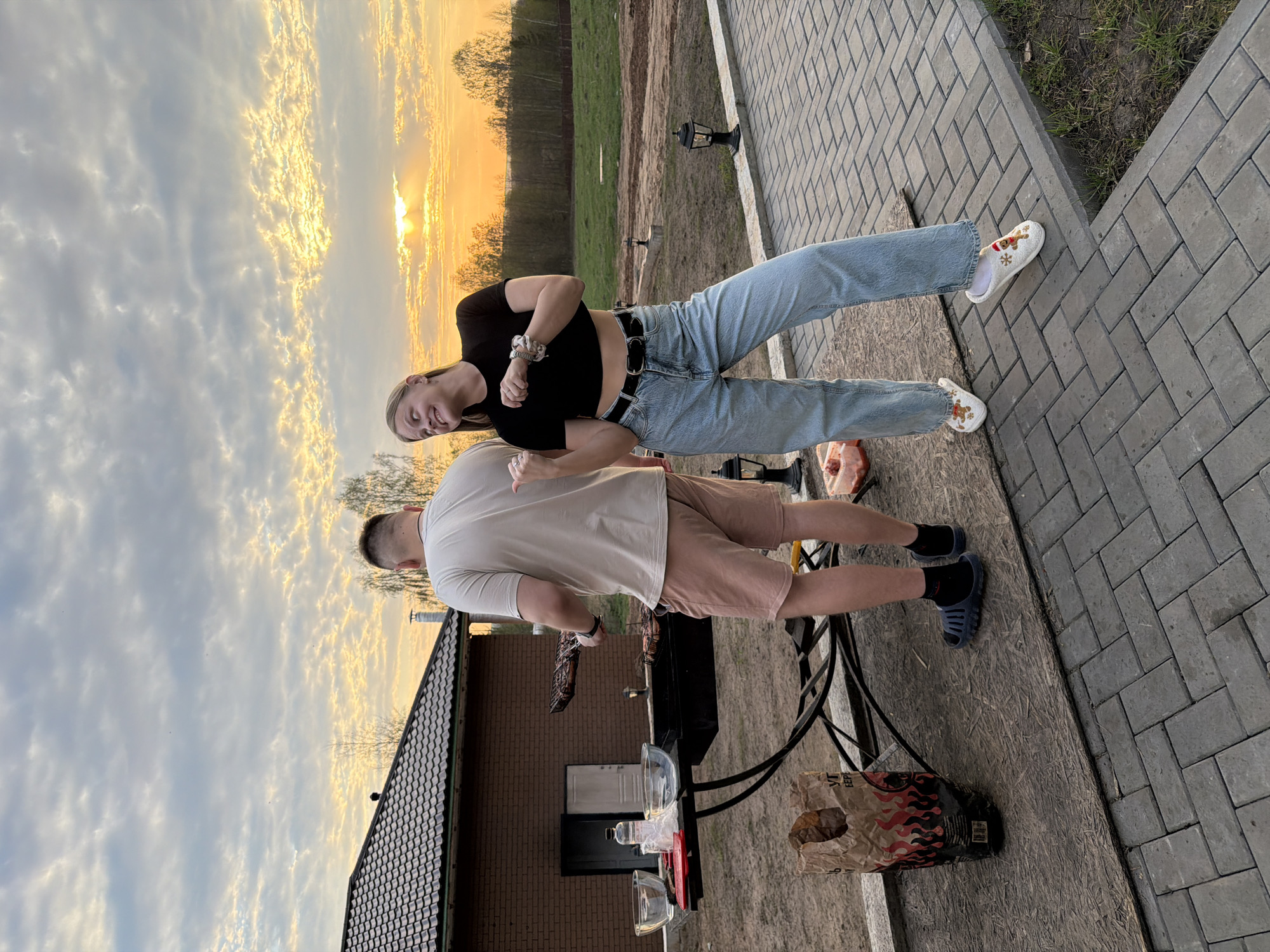 A man and a woman stand on a brick patio near a barbecue grill. The man is grilling food. The woman is standing next to him, smiling. In the background, there is a brick house, grass and trees. The sun is setting. There is a bag of charcoal and a pair of blue slide sandals. The woman is wearing jeans, a black crop top, and white slip-on shoes with embroidered designs. The man is wearing shorts, a t-shirt and socks. The location may be Kamennoye, Russia, based on the prompt.