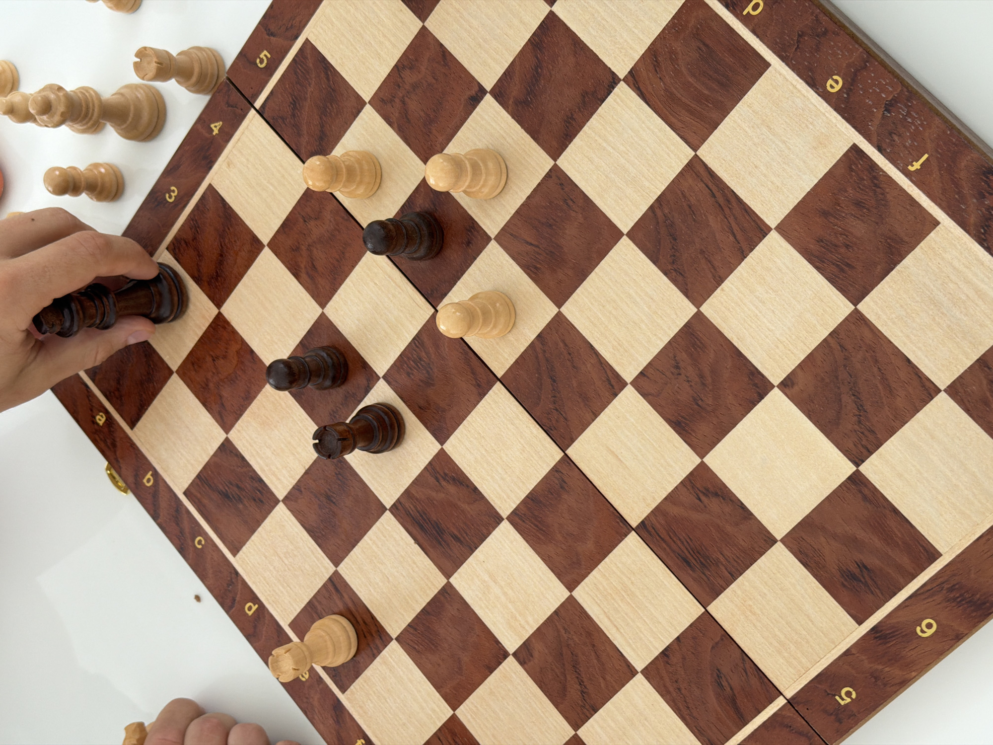 A close-up, high-angle shot captures a game of chess in progress on a wooden chessboard. The board's alternating light and dark wood squares are clearly visible, with a border marked by chess notation letters and numbers. Several chess pieces are scattered across the board: two dark pawns, two light pawns, two dark rooks, and one light rook are identifiable. A hand is visible, mid-action, holding a dark king and about to place it on a square. The background is a clean, white surface, suggesting an indoor setting. There is no information in the image to suggest a location in Thep Krasatti, Thailand.