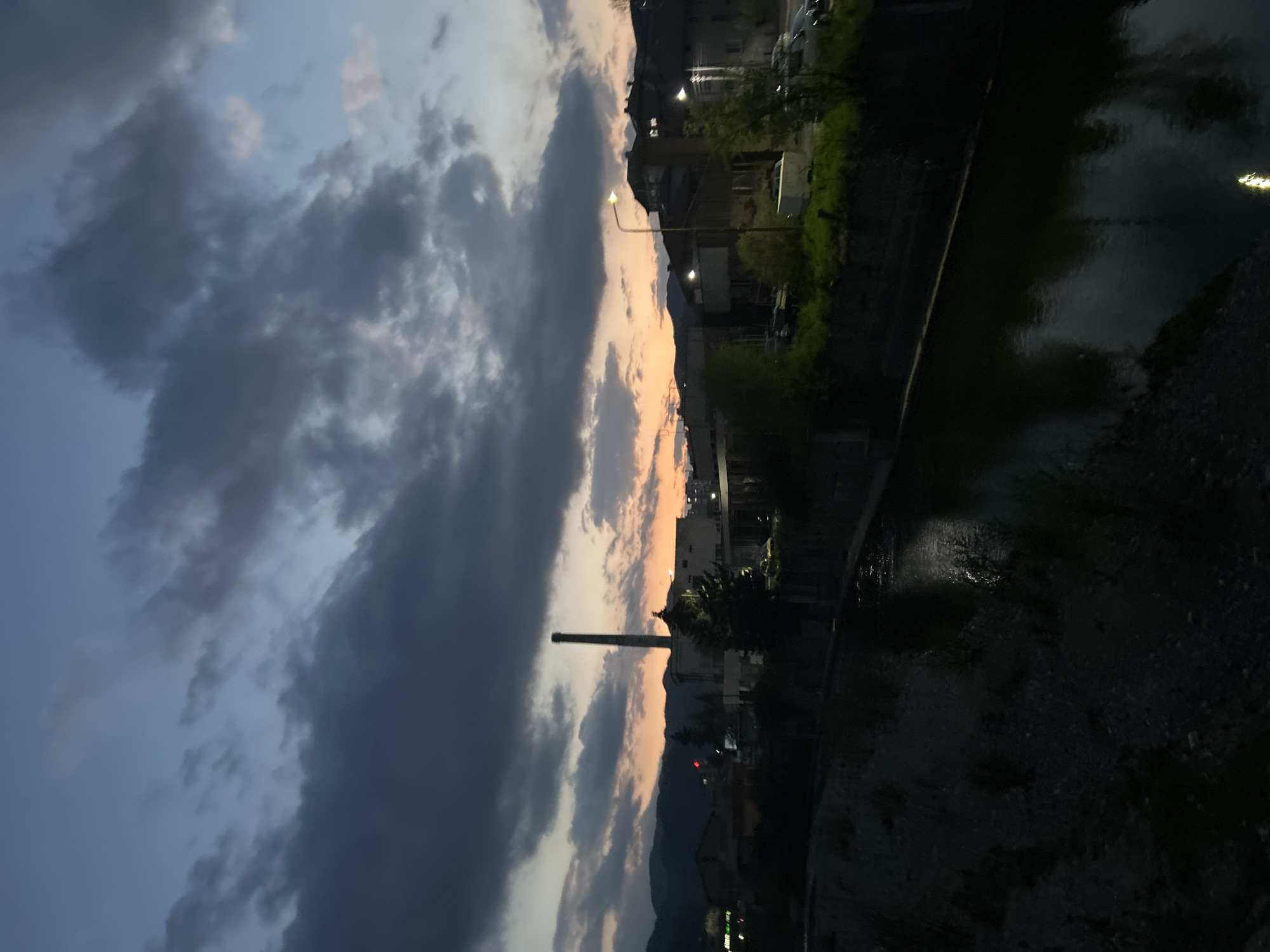 The image captures a twilight scene in what appears to be Zlatograd, Bulgaria. A river or canal runs along the right side of the frame, its surface reflecting the ambient light. The near bank slopes downward and appears to be paved or covered with gravel. Buildings line the far bank, with streetlights illuminating the area. A tall chimney or tower stands out against the sky, which is filled with dramatic clouds in shades of gray, blue, and orange, suggesting either sunrise or sunset. Mountains are visible in the distance. The overall scene gives a sense of urban development meeting natural landscape in a twilight setting.