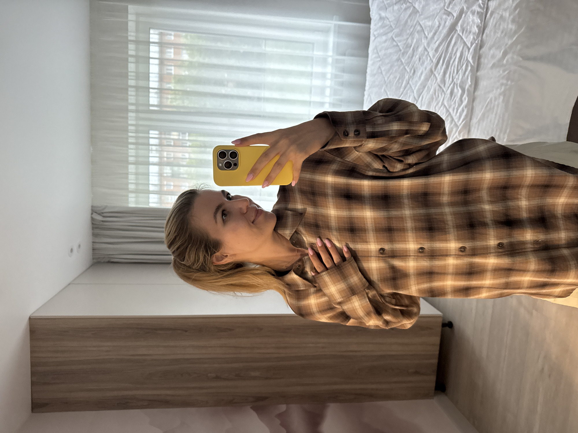 A woman is taking a mirror selfie in a room. She is standing in front of a closet with wooden doors and a white wall. She is wearing a brown plaid shirt and has her hair in a ponytail. She is holding a yellow iPhone in her right hand. There is a white bed with a quilted comforter to her right. A window with sheer curtains is behind the bed, revealing a brick building and trees outside.