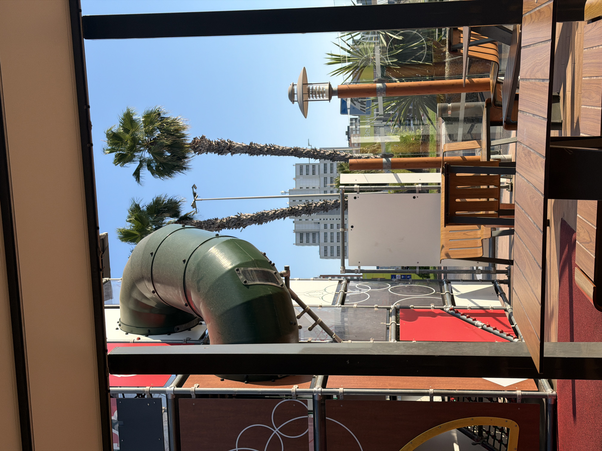 This image depicts an outdoor dining area with a playground structure visible in the background. In the foreground, there are several wooden tables and chairs, some with sunlight casting shadows on them. To the left, part of a red and brown playground structure with a green tube slide is visible. Beyond the playground, two palm trees stand tall against a clear blue sky. Further in the background, several modern buildings can be seen, indicating an urban setting. The scene appears to be taken from inside an establishment, looking out onto an outdoor patio and playground area. The location is identified as Sedaví, Spain. There are no people visible in the image.