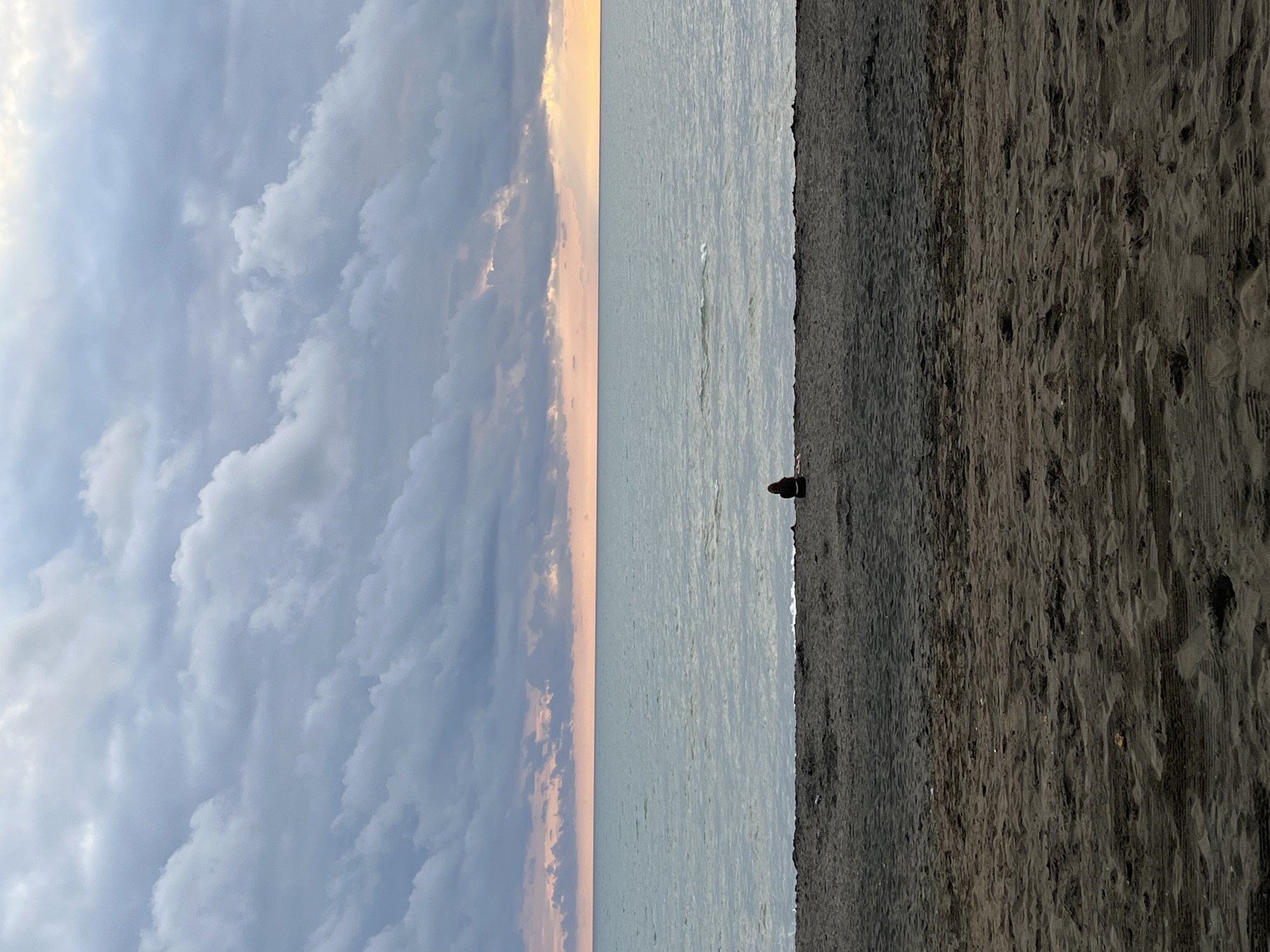 The image shows a serene beach scene in Ureki, Georgia, at dusk. The foreground is dominated by dark, coarse sand, with subtle undulations and traces of previous activity. In the mid-ground, a lone individual, silhouetted against the horizon, sits facing the sea. The sea itself is calm and vast, reflecting the subdued colors of the sky. The horizon line is clearly defined, separating the water from the sky. The sky is filled with a dramatic display of clouds, ranging from dark, heavy masses to lighter, softer formations. A faint band of orange and pink hues suggests the last light of sunset peeking through the clouds near the horizon. The overall atmosphere is one of tranquility and introspection.