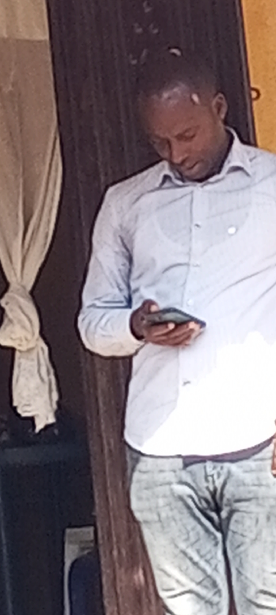 A man stands in front of a doorway in Sabon Tasha, Nigeria, looking at his mobile phone. He wears a light blue, button-down shirt and grey pants. A curtain hangs to his left.