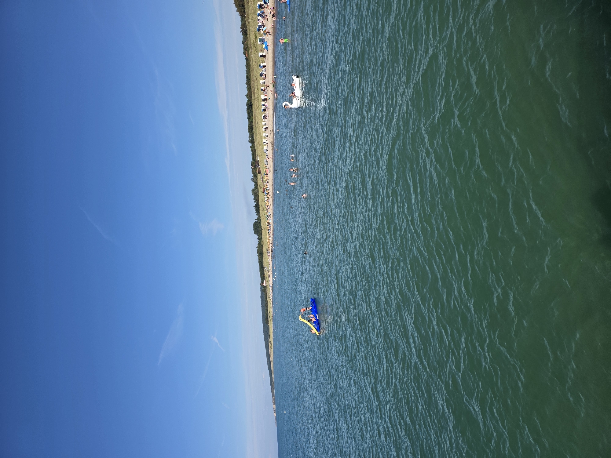 The image shows a sunny day at a beach in Wangels, Germany. The foreground is dominated by the green, wavy water of the sea. In the middle ground, there are several people enjoying the water. One group is on a blue inflatable watercraft with a yellow slide, with at least two individuals on it. Further to the right, a white swan-shaped inflatable is visible, with at least one person on it. Other people are swimming or wading in the sea. The background reveals a sandy beach lined with rows of beach chairs and umbrellas, backed by a grassy dune area and trees. A clear blue sky with a few wispy clouds stretches above.