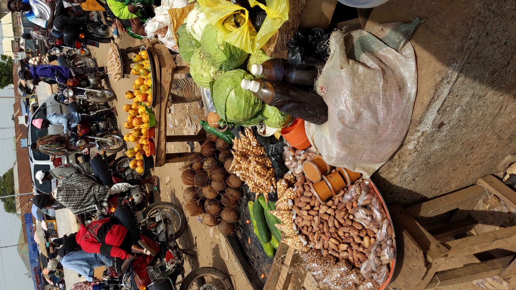 The image depicts a vibrant and bustling outdoor market scene in Ado, Nigeria, under bright daylight. The ground appears to be a mix of dirt and compacted earth.
On the right side, a makeshift stall is laden with a variety of produce and goods. Large, fresh green cabbages are piled high, along with several dark-colored liquids in plastic bottles, possibly palm oil. A large, off-white sack, likely containing bulk goods, is partially visible. Further along the stall, there's a substantial mound of coconuts, several green cucumbers, and numerous small, clear plastic bags filled with shelled peanuts. A large orange tray holds a collection of what appear to be dates or dried fruits, also in small plastic bags, alongside smaller orange containers with a paste-like substance. Another display features ripe yellow mangoes, some individually placed in small, colorful plastic containers.
On the left, the scene is dominated by people and motorcycles, indicating a busy thoroughfare adjacent to the market. Several individuals are seated on motorcycles, either parked or in motion, while others are standing. People are dressed in casual attire, with various head coverings and caps visible. One person wears a red jacket, and another a plaid shirt. In the background, various simple buildings with corrugated metal roofs can be seen, suggesting a semi-urban setting.
The overall activity points to a lively commercial environment with vendors selling goods and people commuting or shopping. The strong sunlight suggests it's daytime, likely morning or early afternoon, with clear weather. No discernible text is clearly visible on any signs, packaging, or vehicles within the frame.