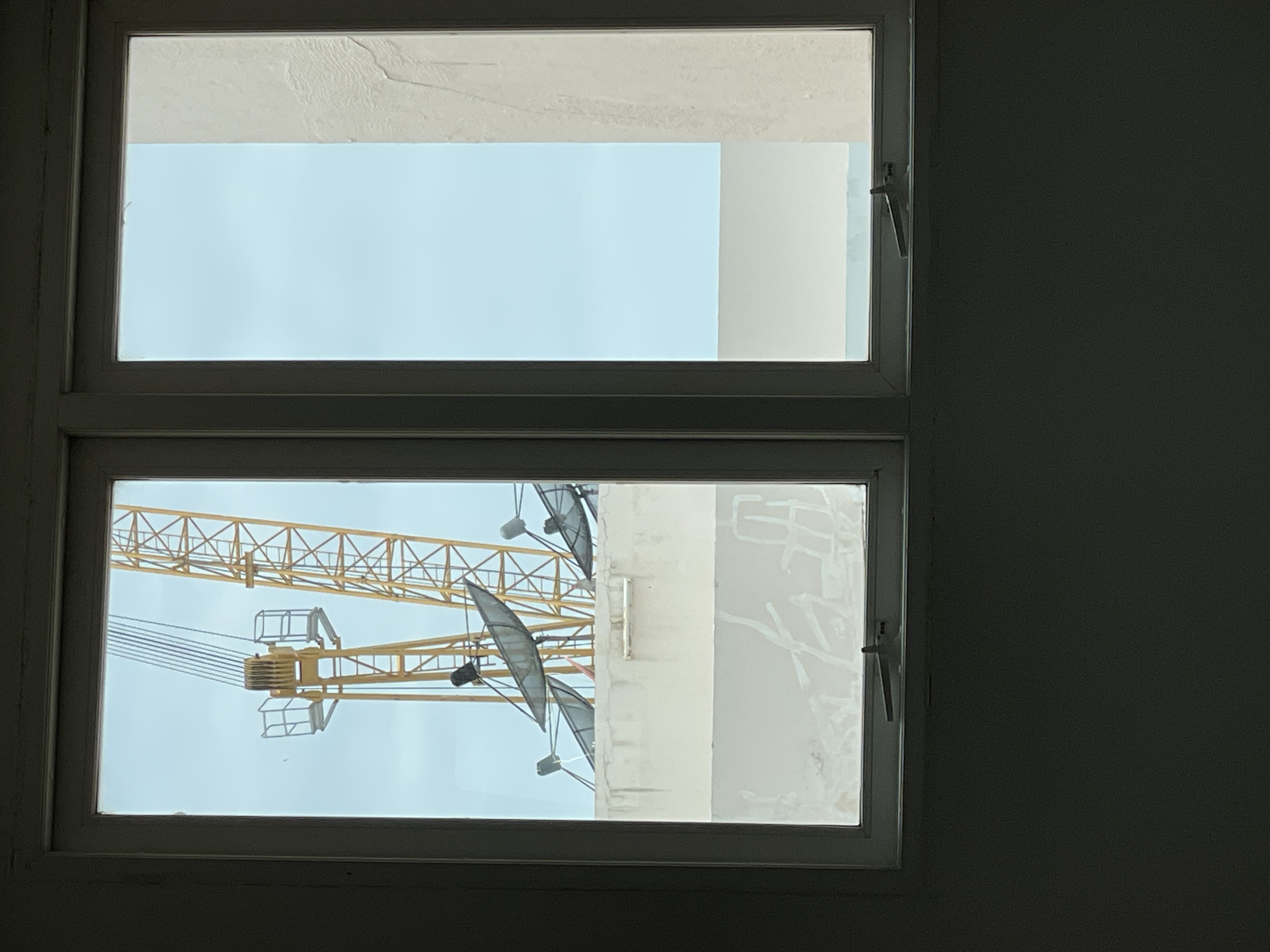 A view through a dual-pane window in Pattaya City, Thailand, reveals a construction scene. In the left pane, a large yellow construction crane with visible cables and a cage-like structure is prominent against a pale blue sky. Below the crane, several satellite dishes are mounted on a white rooftop structure. The right pane shows a continuation of the pale blue sky and a textured, off-white building facade. The window frame is white, and the interior foreground is dark.