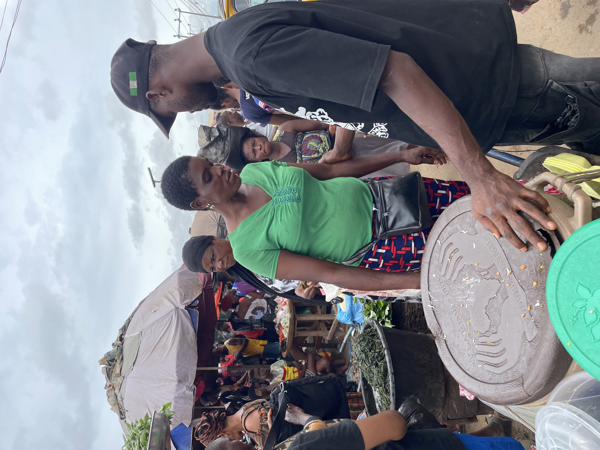 This is a medium shot of a busy outdoor market scene with a cloudy sky overhead. In the foreground, a man wearing a black t-shirt and black jeans, a black baseball cap with the Nigerian flag on it, stands facing to the right, his left arm outstretched. To his left, a woman in a green t-shirt and a red, white, and blue patterned skirt stands with her left hand near her hip and her right hand holding something green and leafy. Behind them, other people are visible, including a woman wearing a black headscarf and a black and white patterned dress, looking to the left. A large, dark brown circular lid with an embossed design is in the lower center of the frame, with a person's hands touching it. There are also large black tubs filled with leafy green vegetables in the left foreground. The market appears to be selling produce, with various stalls and umbrellas visible in the background. The overall impression is one of a vibrant, active marketplace.