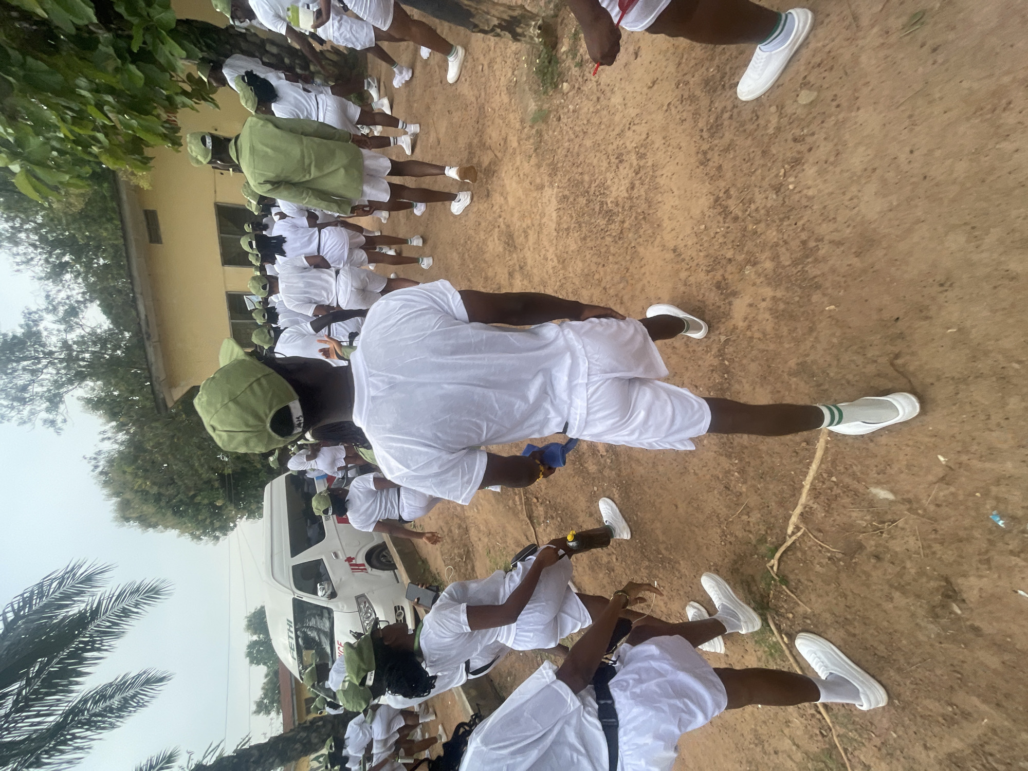 A group of young adults, dressed in white t-shirts, white shorts, and green caps, are gathered outdoors on a dirt path. Many are facing away from the camera, walking in the same direction. A white van, labeled "FETHI" and with a red cross symbol, is parked in the background. The scene appears to be at a camp or training facility, given the uniform attire and the presence of what could be a medical vehicle. The environment is somewhat unkempt, with visible tree roots and scattered debris on the ground. The weather seems overcast. The group's attire suggests they might be participants in a youth service program or a similar organized event. This image captures a moment in a communal gathering, possibly on a field day or during a period of organized activity.