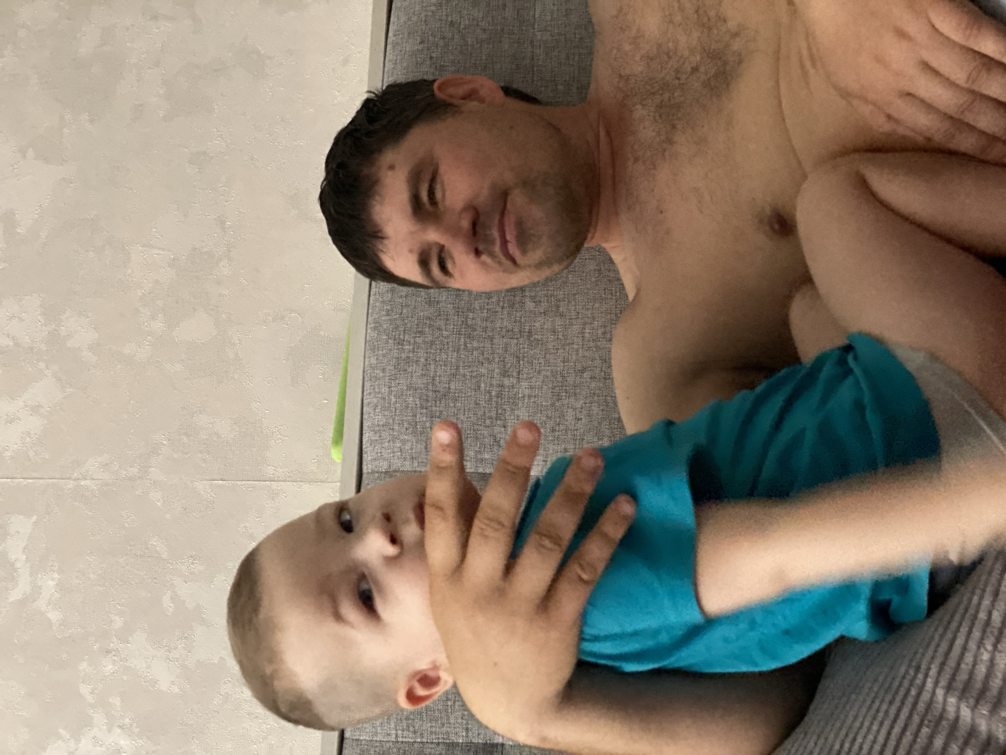 A man and a young child are lying on a grey sofa. The man is shirtless and has dark hair. The child, wearing a teal shirt, is positioned on the man's chest with their hand partially obscuring their face. The location is suggested to be Sochi, Russia. The environment appears to be an interior setting with a textured wall visible in the background.