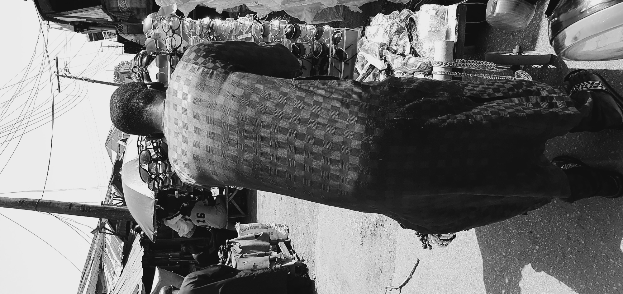 In a black and white outdoor scene, an adult male is captured from behind, standing in what appears to be a marketplace in Bukuru, Nigeria. He is wearing a patterned long shirt or tunic and dark trousers. His hair is short. In front of him and to his right, there are stacks of sunglasses displayed on stands. Further to the right, there are several plastic bags filled with items, and a chain with a pendant hangs from a cart. To the left of the frame, another person is partially visible, walking away, and behind them, a market stall with a thatched roof is visible, along with what appears to be a sack of flour. In the background, several structures with corrugated metal roofs are visible, and a utility pole with numerous wires stretches across the sky. The ground is a dirt or gravel surface. The lighting suggests it is daytime.