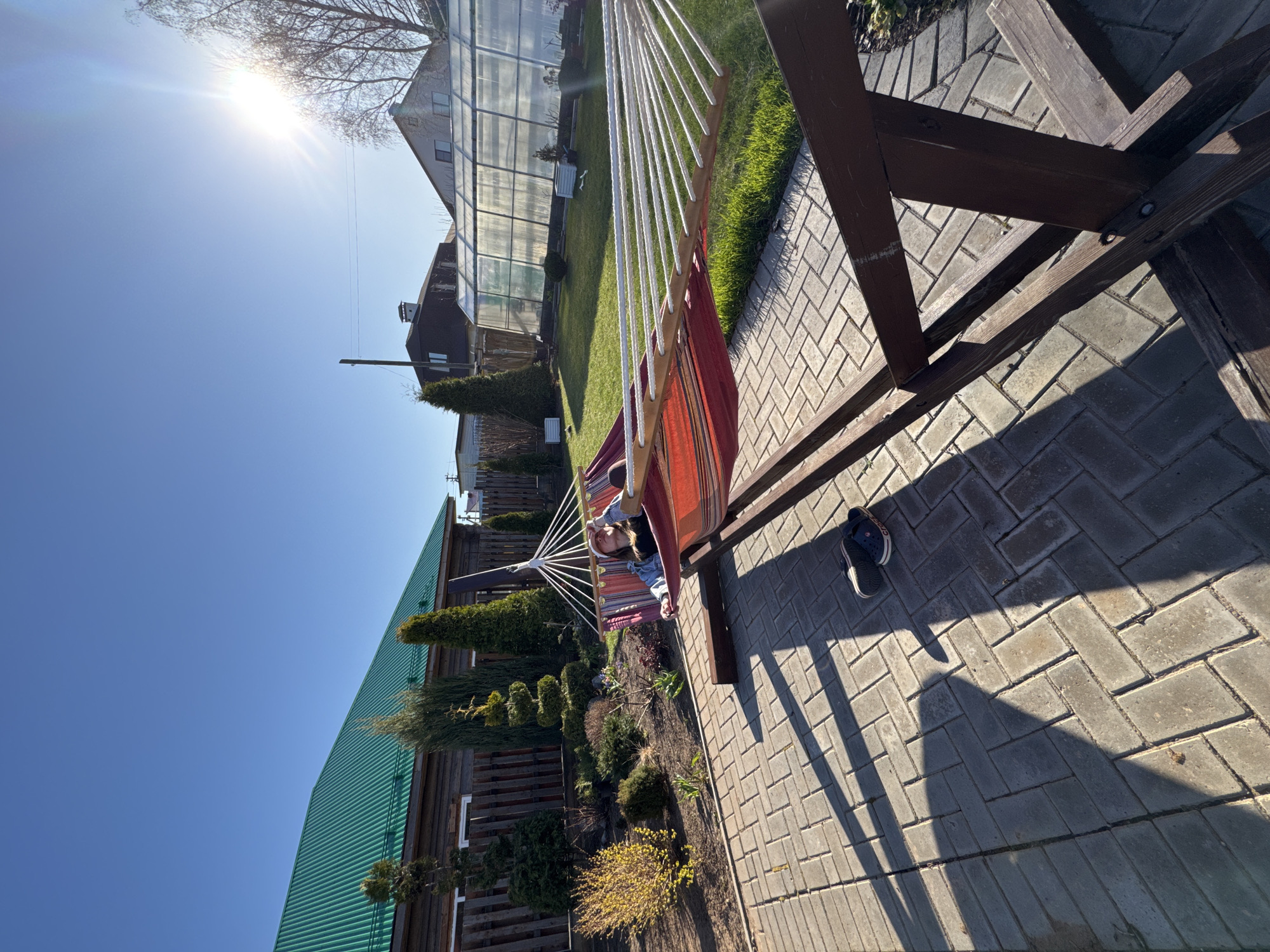 The image depicts an outdoor scene in Прикамский, Russia, featuring a hammock suspended between a wooden frame. A person is lying in the hammock. The ground is paved with gray bricks, and a pair of dark blue Crocs shoes are visible. In the background, a wooden building with a green roof, evergreen shrubs, a garden, and a greenhouse are visible. The sky is blue with a bright sun.
