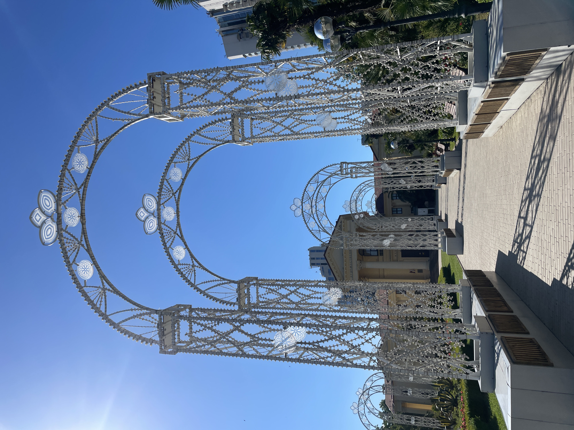 The image depicts a bright, sunny day in Sochi, Russia, likely in a public park or promenade, characterized by a series of large, ornate white arches. These arches are intricately constructed with a metallic lattice framework adorned with delicate, snowflake-like decorations and small lights. The arches form a pathway leading towards a classical-looking building with columns, suggesting a cultural or governmental establishment.
The foreground features a paved walkway with a light brown, brick-like pattern, bordered by raised planters with wooden benches integrated into their structure. Lush green grass and some exotic plants, including what appear to be agave plants, are visible in the planters. The arches are positioned along the pathway, with the largest and most prominent ones closest to the viewer.
In the midground, the pathway continues between more arches, leading towards the central building. Palm trees and other tropical vegetation frame the scene on the right side, with a modern building visible behind them. The sky is a clear, vibrant blue, indicating fair weather. There are no people visible in the image. The overall impression is one of a well-maintained, decorative public space, likely designed for pedestrian use and aesthetic appeal, possibly for events or seasonal decorations.