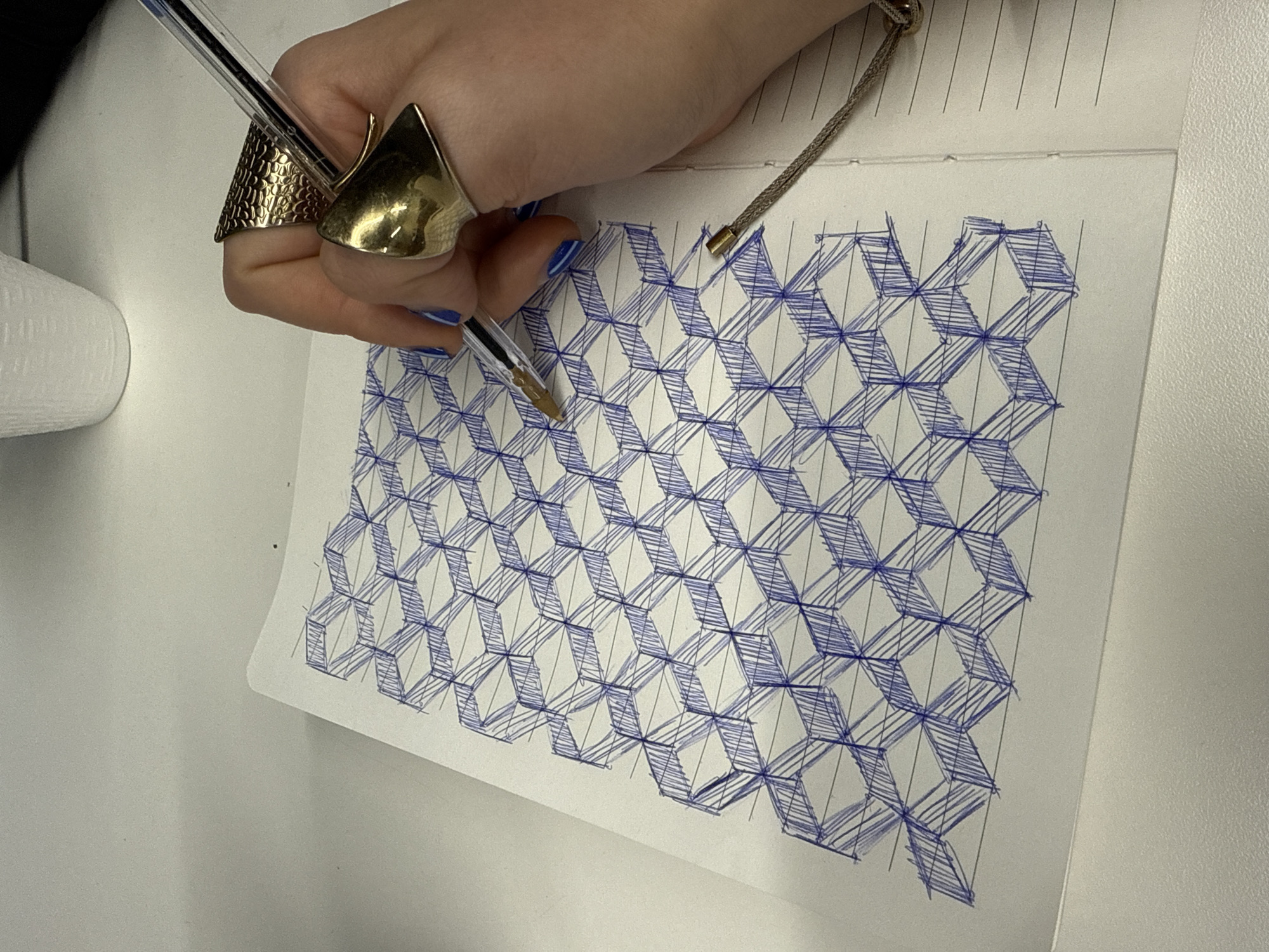 A medium shot, taken from an elevated angle, shows a hand drawing a geometric pattern on a piece of paper. The hand is holding a clear ballpoint pen, with a gold-colored point visible. The hand is adorned with two large, gold-colored rings; one has a textured surface, while the other has a smooth, pointed shape. The fingernails of the hand are painted blue.
The paper contains a pattern of interlocking blue-inked, rhomboid shapes that create an optical illusion of cubes. The pattern is not perfectly executed; there are gaps, overlaps, and inconsistencies in the lines. The paper rests on a lined notebook. A gold chain is partially visible at the top of the notebook. To the left of the paper, part of a white, cylindrical object is visible. To the right of the paper, a white, flat surface is visible.