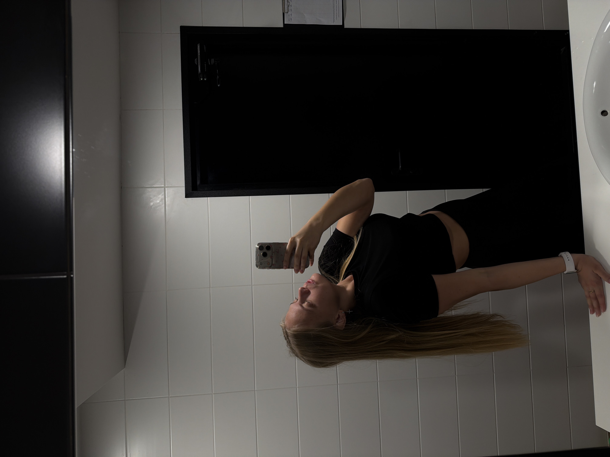 A young woman with long blonde hair, dressed in a black top and black pants, is depicted taking a mirror selfie in an indoor, tiled setting, likely a bathroom or changing room. She holds a silver smartphone, possibly an iPhone, in her right hand, its screen reflecting her face as she poses. On her left wrist, she wears a white smartwatch, and a ring is visible on her right hand. Small cross earrings can be seen on her ears.
The background features white rectangular wall tiles. To the right, a white sink or basin is partially visible. A large, dark rectangular object, resembling a cabinet door or window frame, is prominent in the central background. Above this dark object, a clipboard is mounted, bearing faint, illegible text. The lighting is artificial, illuminating the scene. The woman's expression is neutral, focused on her reflection and the selfie-taking process. This scene is located in Izhevsk, Russia.