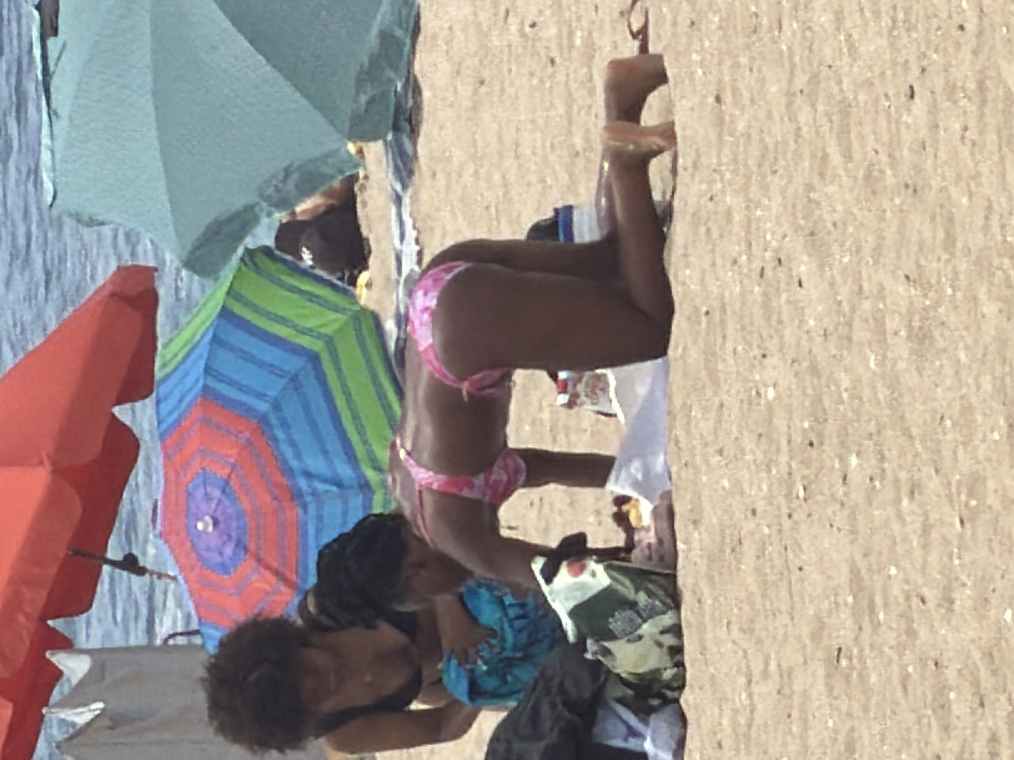 Two women are on a sandy beach. The woman in the foreground is on her hands and knees, wearing a pink and white patterned bikini. She is leaning forward, reaching towards a camouflage patterned bag and a white towel on the sand. Her legs are bent, and her feet are visible. The woman in the background is sitting down, wearing a black bikini top. She has dark, curly hair and is holding a blue object in her lap. Behind them, various beach umbrellas are visible, including a red one, a large blue and green striped one, and a light blue one. The ocean is visible in the background. The image is a photograph taken on a bright day.
