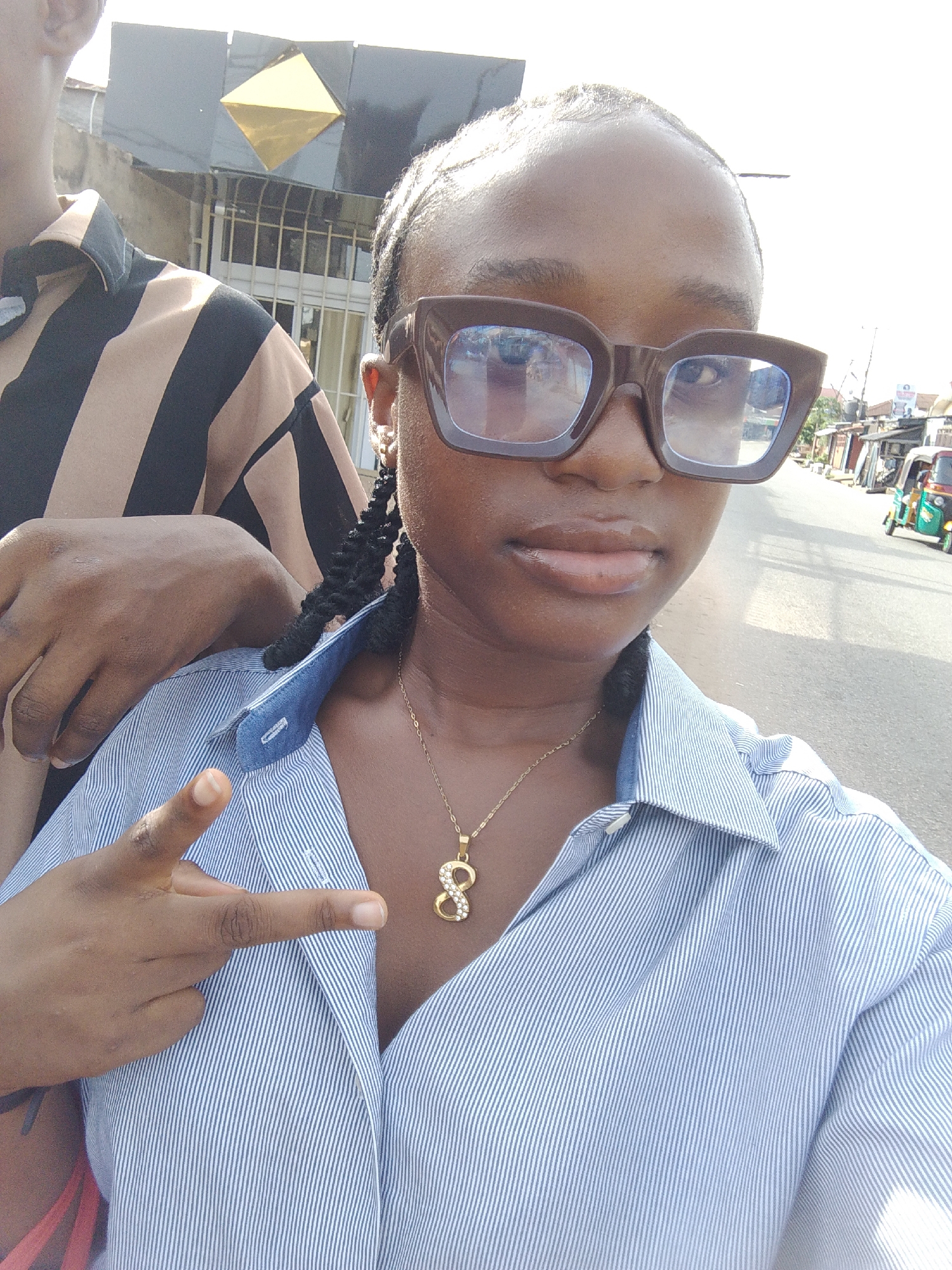 A close-up selfie-style outdoor photograph captures two individuals on a street in Ohokobe Ndume, Nigeria, under bright daylight. The primary subject, a person with dark skin, occupies the foreground, wearing large, dark-framed rectangular eyeglasses that obscure most of their eyes. Their lips, with a slight sheen, are visible, and their hair appears to be styled in braids. They are dressed in a light blue and white pinstriped collared button-up shirt, partially unbuttoned, revealing a gold chain with a diamond-studded pendant resembling the number '8' or an infinity symbol. Their left hand is raised, forming a "peace" or "V" sign with the index and middle fingers.
To the left of the main subject, another person is partially visible, clad in a light brown and black striped short-sleeved collared shirt. Their arm rests casually on the main subject's shoulder.
The background reveals a sunlit street scene characteristic of a local urban or suburban area. Buildings line the paved road; one prominent dark-colored building directly behind the partial subject features a golden, diamond-shaped architectural embellishment. Further down the street on the right, a green and yellow three-wheeled auto-rickshaw (tuk-tuk) is visible, alongside other indistinct structures. The bright, direct sunlight suggests a clear, sunny day, likely late morning or afternoon. The overall impression is casual and relaxed. No discernible text is visible on signs or clothing.