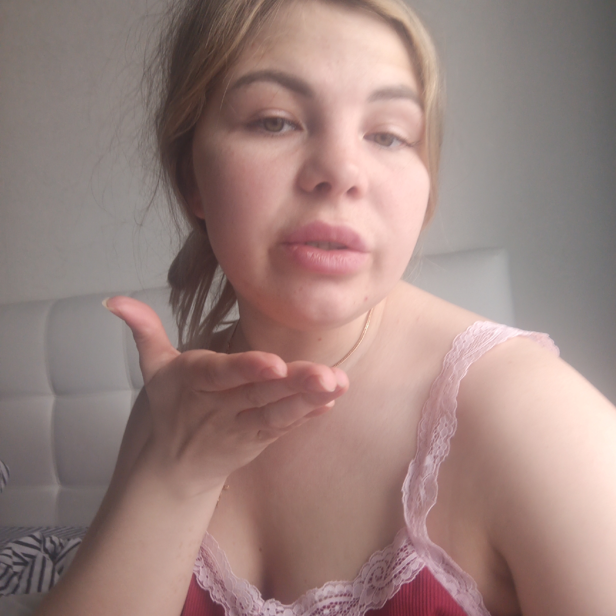 A young woman with light brown hair is captured in a close-up, chest-up shot, looking directly at the camera. She has her lips puckered, as if blowing a kiss, and her right hand is cupped and extended forward to complete the gesture. Her eyes, which appear brown or hazel, are open and engaging. She wears a reddish or maroon ribbed tank top with light pink lace trim along the neckline and straps, and a thin gold-colored chain necklace is visible around her neck.
The setting appears to be indoors, likely a bedroom, indicated by a white, padded headboard visible behind her. Striped bedding in black and white can be seen in the bottom left corner. The lighting is soft and bright, suggesting natural light from a window. The woman's expression conveys a sense of playfulness or affection. No visible text is present in the image.