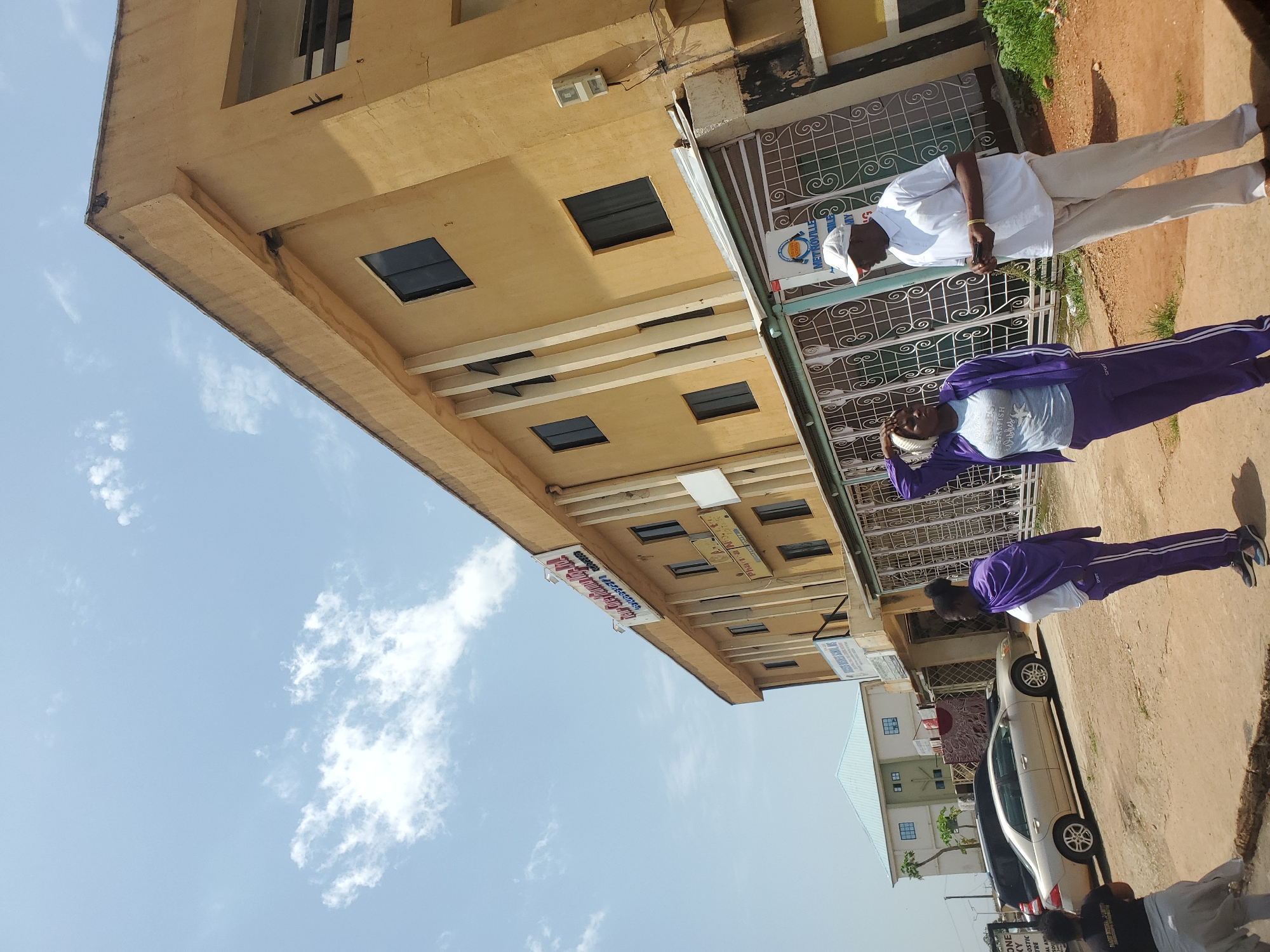 The image depicts two individuals in an outdoor urban setting, likely Jos, Nigeria. The primary subjects are a man and a woman, both appearing to be of African descent. The man, standing upright, is dressed in a white short-sleeved shirt, light-colored trousers, and a white cap. He is holding a mobile phone in his right hand. The woman, positioned slightly further away and to the left, is wearing a purple tracksuit over a light-colored t-shirt and white headscarf. She has her right hand raised, touching her head.
They are standing on a dirt path or road beside a two-story building with a yellow facade. The building has several square windows and an overhang. There are also metal security bars on some of the windows and doorways. Faintly visible on the building is a sign that appears to have text, though it is difficult to decipher clearly. In the background, there are other buildings, including one with a light blue roof, and a silver sedan car parked on the right side of the image. The sky above is a clear blue with scattered white clouds, suggesting it is daytime with fair weather. The overall scene appears to be a casual street view.