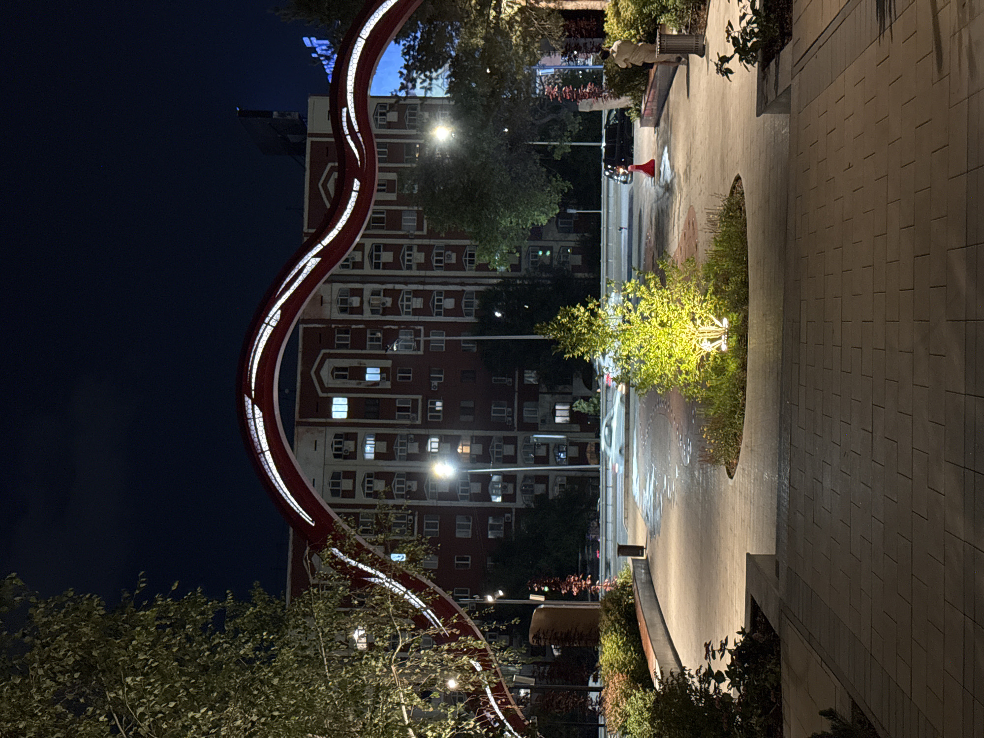 The image depicts a nighttime view of a city square in Almaty, Kazakhstan, featuring a prominent red, curved archway with integrated lighting. The arch frames a multistory brick building with numerous windows, many of which are illuminated, suggesting residential or office use. In the foreground, a paved walkway leads into the square, flanked by landscaped areas with trees and bushes. A small, brightly lit tree is situated in the center of the square. The ground in the square appears wet, reflecting the ambient light. A white car is visible in motion on the street behind the square, and a black car and an orange traffic cone are present near the edge of the square. A person in light-colored clothing stands to the right of the frame, near a trash receptacle. The overall scene conveys an urban, modern aesthetic with an emphasis on nighttime illumination and architectural design.