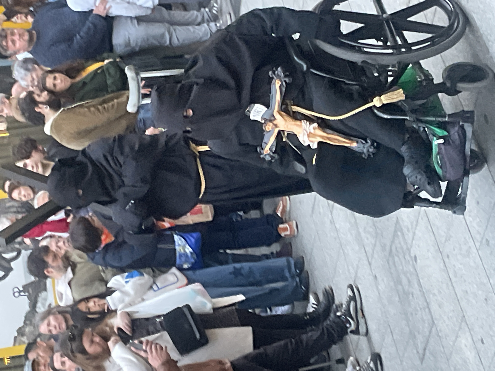 A person wearing a black hooded robe and seated in a wheelchair is visible in a crowd of people. A crucifix is prominently displayed on their robe. They also have a yellow rope belt. The location is likely Barcelona, Spain. The person is stationary amidst the crowd. The environment is an outdoor, public area with tiled flooring.