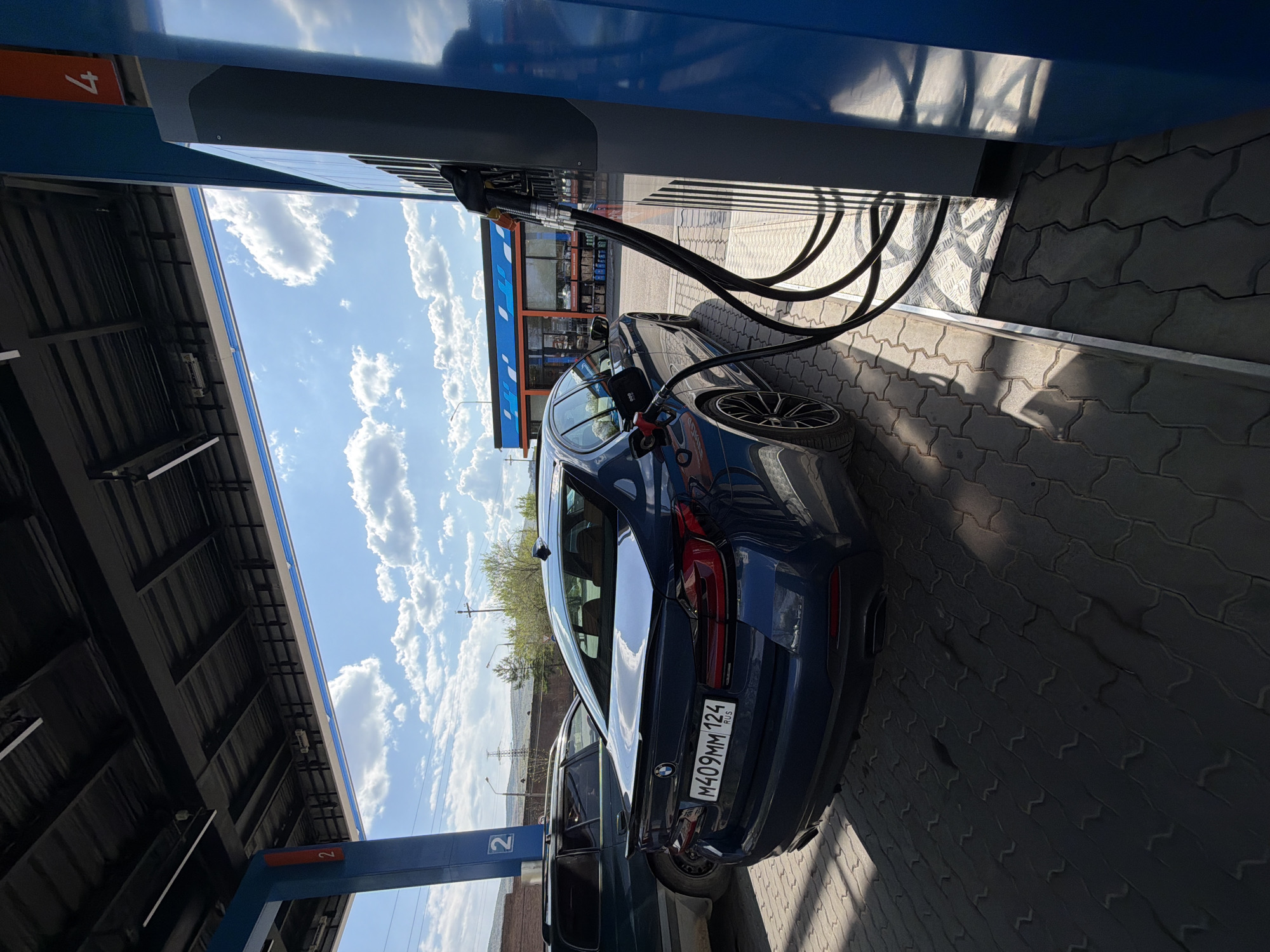 A dark blue BMW is parked at a gas station in Krasnoyarsk, Russia. The car is positioned next to a gas pump, and a black fuel hose is inserted into the car's fuel tank. The license plate number is M409MM124. The gas station has a canopy overhead, and the sky is visible in the background with scattered clouds. The gas station's exterior has a blue and white color scheme. The ground is paved with interlocking concrete pavers. Another car is partially visible adjacent to the BMW. A pole labeled "2" is visible in the foreground, and a pole labeled "4" is also visible. The scene is brightly lit, likely due to sunlight.