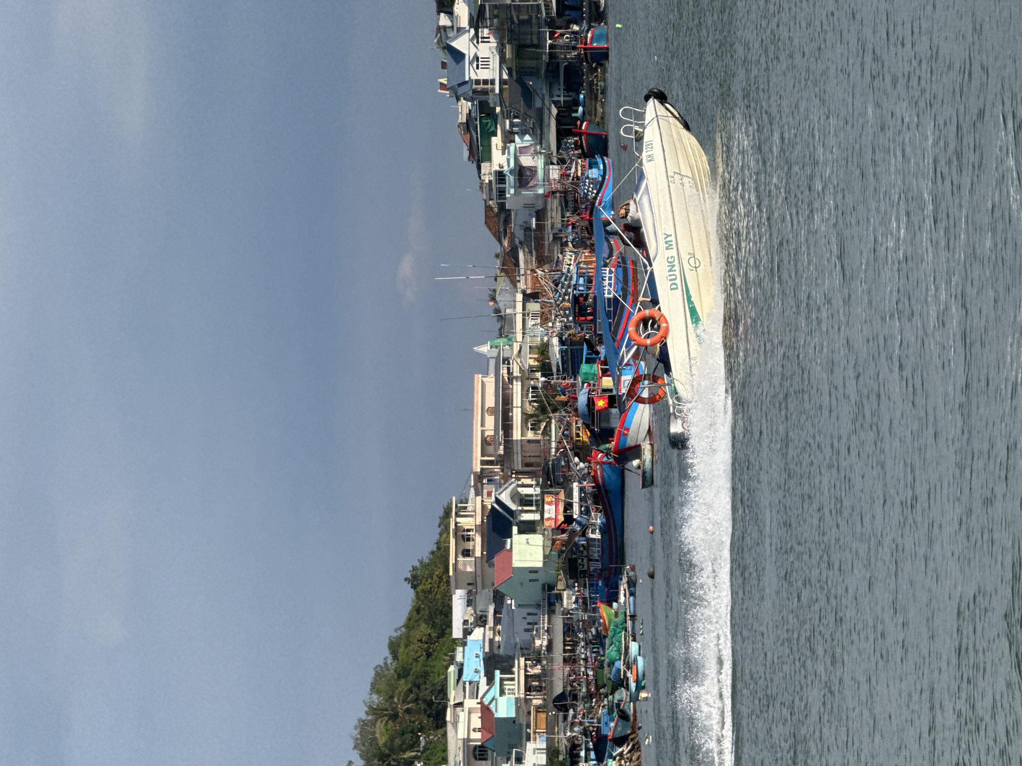 In Nha Trang Ward, Vietnam, a bright white speedboat, emblazoned with "DŨNG MY" and the registration number "KH 1281," cuts through the water, leaving a dramatic white wake. The boat, carrying at least two individuals, is angled towards the right of the frame, its bow slightly lifted. In the background, a bustling harbor teems with numerous fishing vessels of varying sizes and colors, predominantly blues and reds, many adorned with flags. The boats are docked closely together, creating a dense maritime landscape. Beyond the harbor, a hillside is densely packed with buildings exhibiting a range of architectural styles and colors, from pastel blues and greens to whites and yellows, with tiled roofs. The sky above is overcast, with a patch of lighter clouds suggesting the presence of sun. The water's surface is choppy, reflecting the dynamic movement of the speedboat and the ambient light.