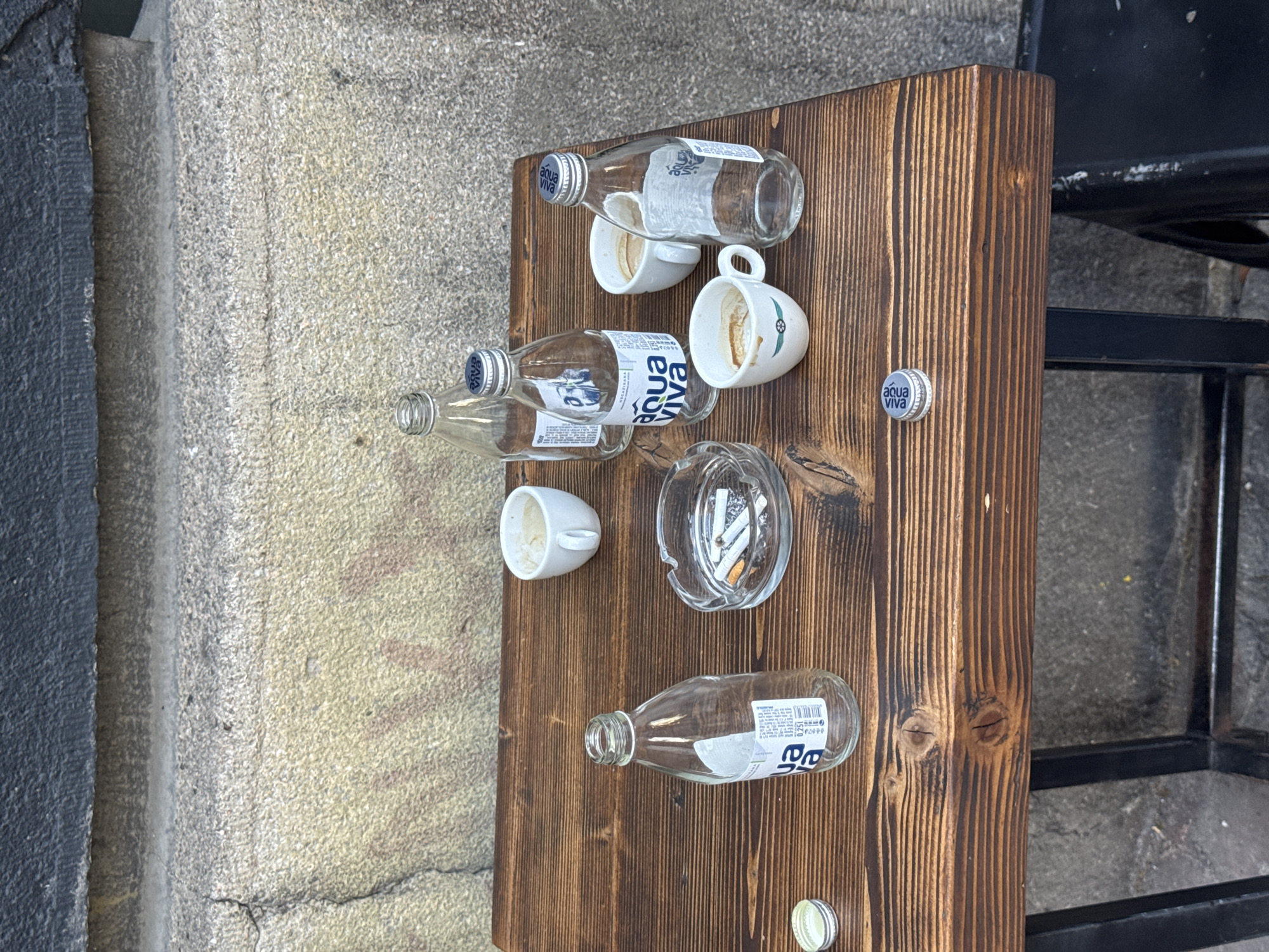 The image shows a brown wooden plank propped up against a wall in an outdoor setting, possibly Belgrade, Serbia, judging by the water bottle brand "Aqua Viva." The plank is littered with discarded items: three empty "Aqua Viva" glass water bottles, two used coffee cups, and a glass ashtray filled with cigarette butts. One "Aqua Viva" bottle cap is also on the board. The plank appears to be used as a makeshift side table, next to a metal chair. The background includes a textured wall and a glimpse of the urban environment.