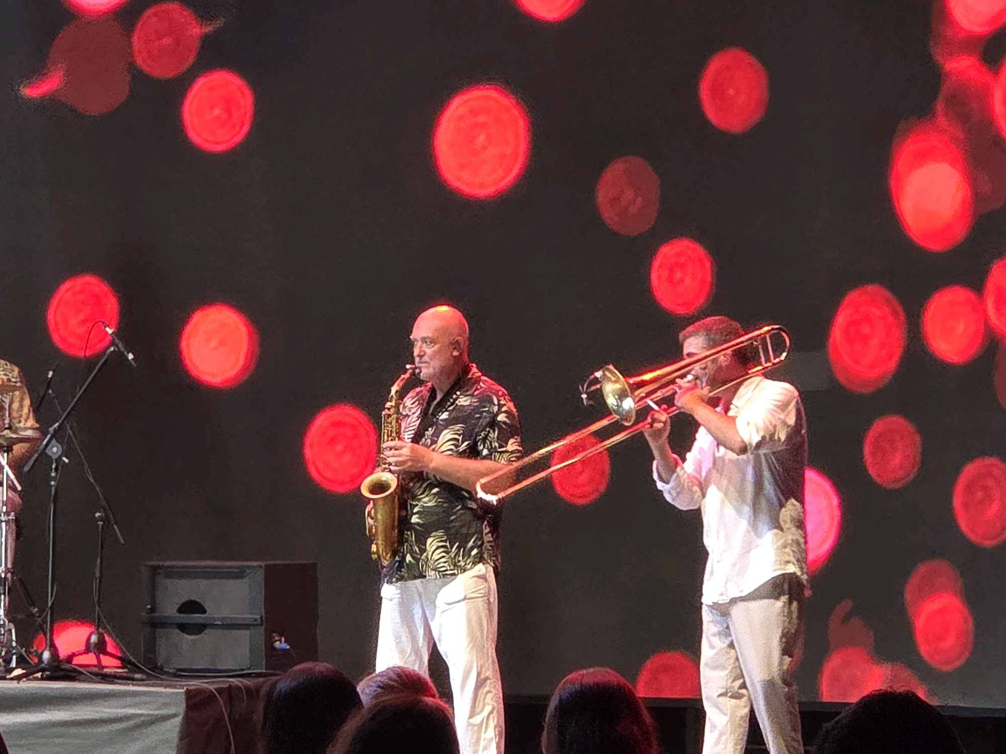 Two musicians are performing on a stage in front of a dark background with large, blurry red circles. A bald man on the left is playing a saxophone. He is wearing a black shirt with a tropical leaf pattern and white pants. To his right, another man is playing a trombone. He is wearing a white shirt and light-colored pants. The stage has a speaker on the left side. The lighting is dim, with the red circles creating a vibrant backdrop. The location is specified as Bodrum, Turkey.