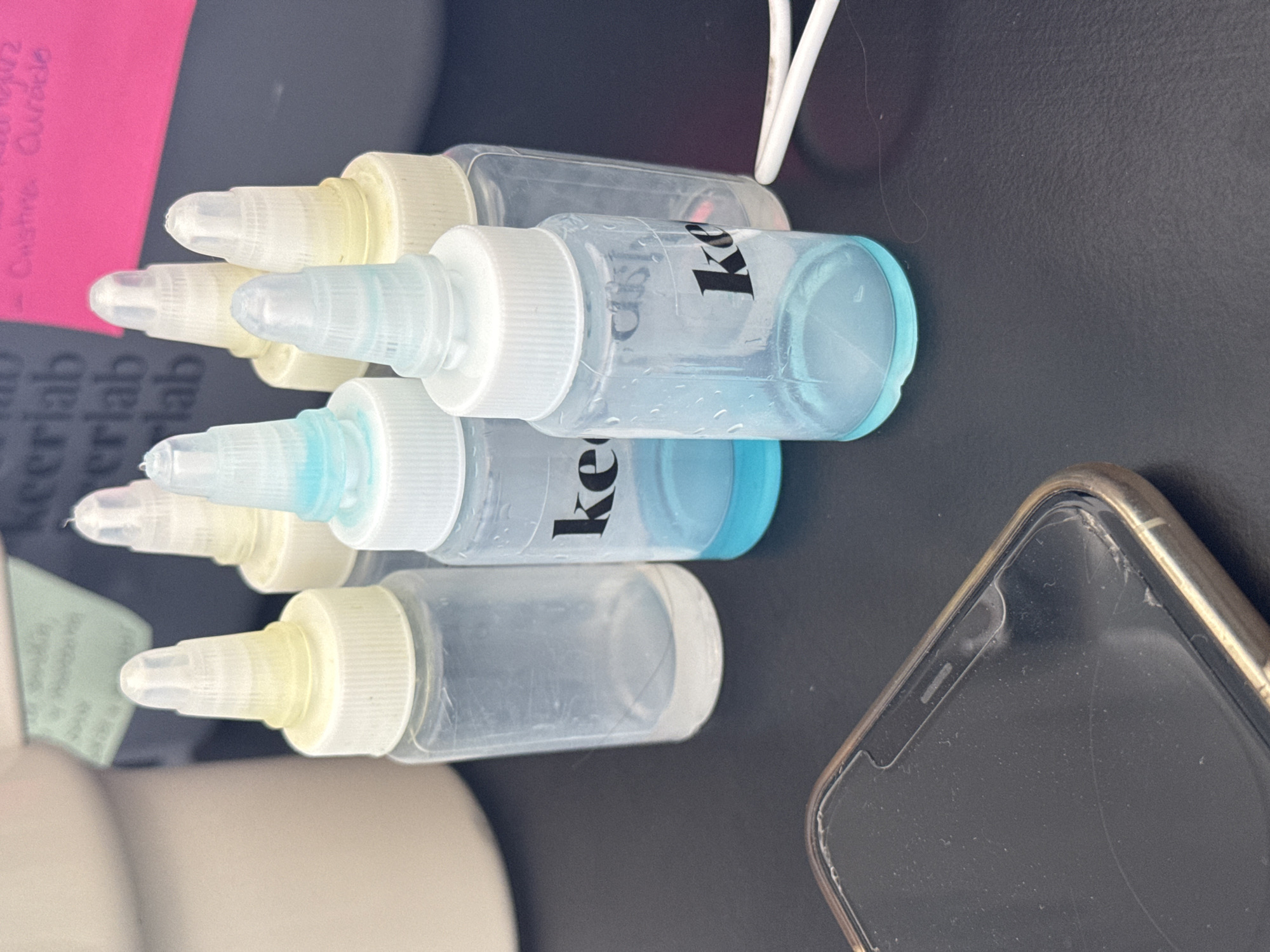 The image shows a close-up view of several small, clear plastic bottles with white caps, lying horizontally on a dark surface. The bottles appear to contain varying amounts of liquid, some clear and some with a blue tint. "Keerlab" is printed in black on at least two of the bottles. A partially visible smartphone, with a cracked screen protector, is in the lower right corner of the image. In the background, there are pink and green sticky notes. The location, San Bernardino Tlaxcalancingo, Mexico, is not discernible from the visual elements of the image.