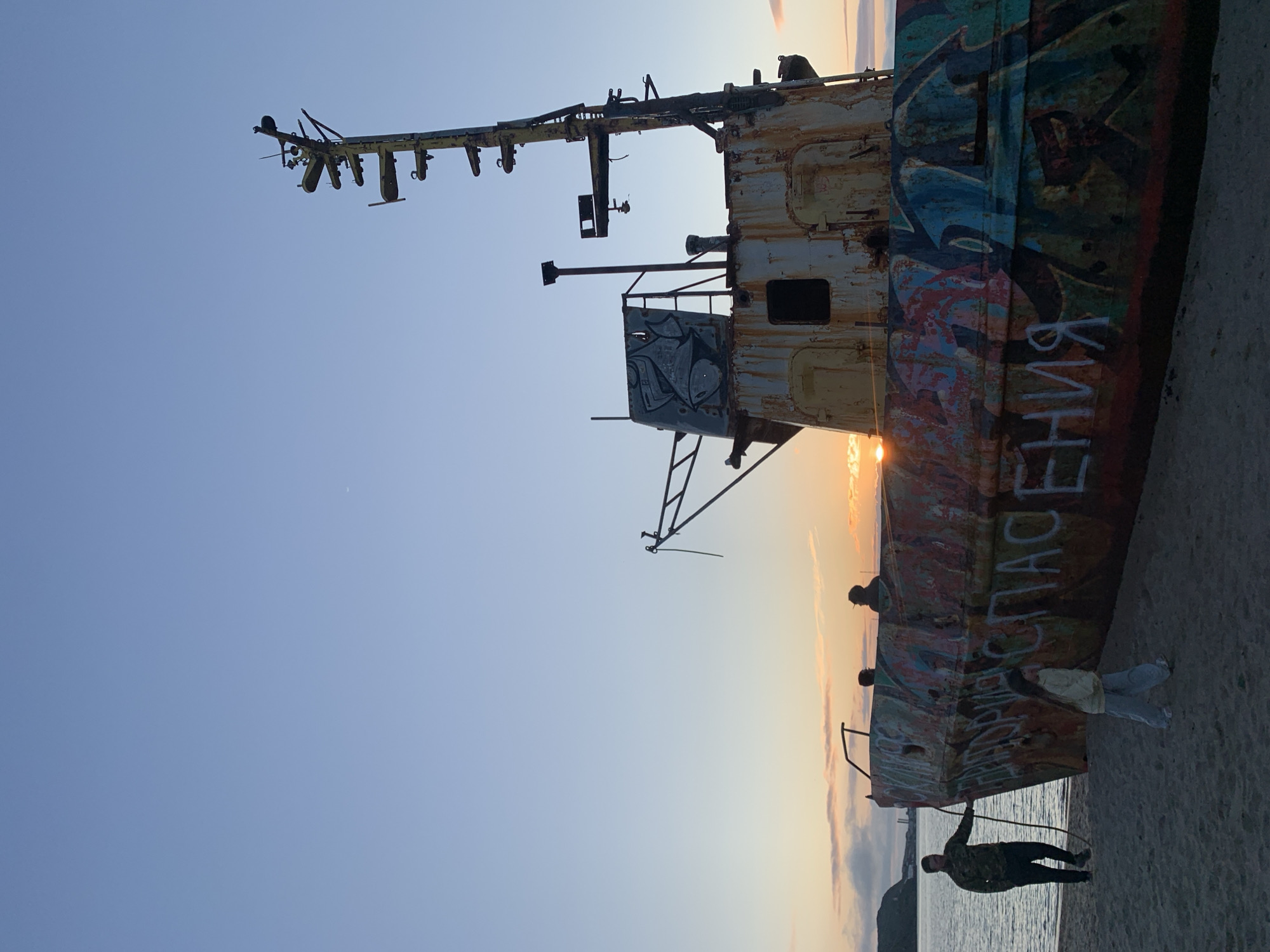 This image depicts a beached, derelict ship on a sandy shore in Teriberka, Russia, during sunset. The ship, heavily vandalized with colorful graffiti, has a rusty and weathered appearance. The sky displays a gradient of blue to pale orange, indicative of twilight. In the foreground, a man in camouflage clothing is standing near the ship, holding a rope. A young girl in casual attire is walking away from the ship towards the viewer. In the background, a few other silhouetted figures can be seen near or on the ship. The sea is calm and to the left of the ship, with a distant coastline visible. The overall scene conveys a sense of abandonment and decay against a backdrop of natural beauty.