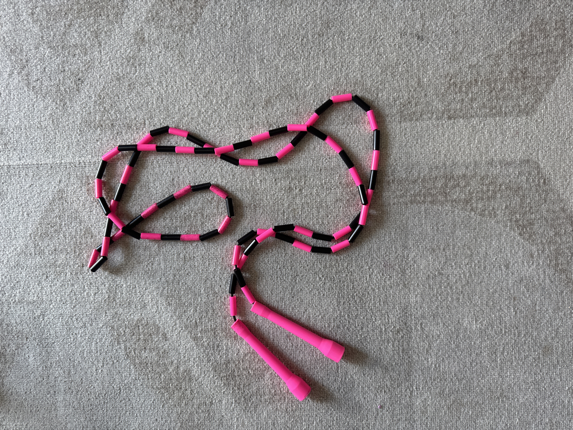 A beaded jump rope with pink handles and a cord alternating between pink and black beads lies on a textured, light gray surface. The jump rope is coiled loosely, forming loops and curves, with the handles positioned towards the bottom right of the frame. The beads appear cylindrical and are threaded together to create the rope. The surface beneath the jump rope resembles carpet or a textured fabric. The lighting is even, suggesting an indoor setting with diffused light, possibly natural light from a window or overhead artificial lighting. No people are visible in the image, and there is no text. The overall impression is that of an object placed on a neutral background, ready for use or storage.