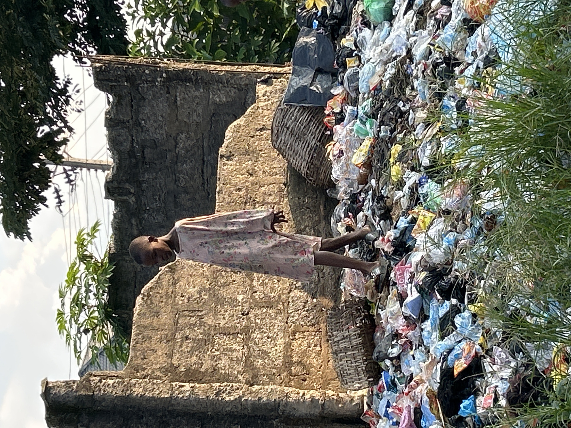 The image depicts a young person in a light-colored dress with a floral pattern, standing on a stone ledge. The person is looking downwards and to their right. Behind the individual, a large pile of garbage is visible, composed of various plastic bags, wrappers, and other debris. A woven basket is partially obscured by the trash. The background shows lush green foliage and a blue sky, suggesting an outdoor setting. The stone structure appears weathered and aged. The lighting suggests it is daytime, possibly late morning or early afternoon, given the shadows. The scene is set in Ihiagwa, Nigeria, a location known for its vibrant culture and sometimes challenging environmental conditions. The overall mood of the image could be interpreted as somber, highlighting the contrast between the natural environment and the human-made waste. There is no visible text in the image.