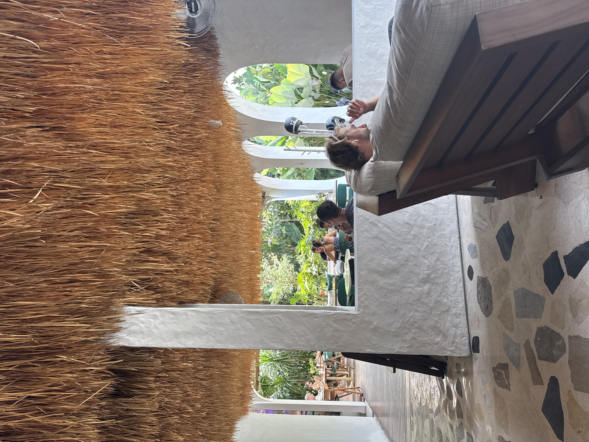 The image, taken in North Kuta, Indonesia, shows an outdoor restaurant or cafe setting with a thatched roof on the left side of the frame. A white, arched wall separates the covered seating area from an open-air section filled with lush green plants and foliage. Inside the covered area, a fair-skinned man is reclined on a wooden chaise lounge with light-colored cushions. His hair is brown. Through the arches, other people are seen seated at tables, seemingly enjoying a meal. The right side of the image features a decorated floor with a mix of light and dark stones. A wooden chaise lounge is shown in the upper right corner. The setting appears to be a blend of indoor and outdoor spaces.