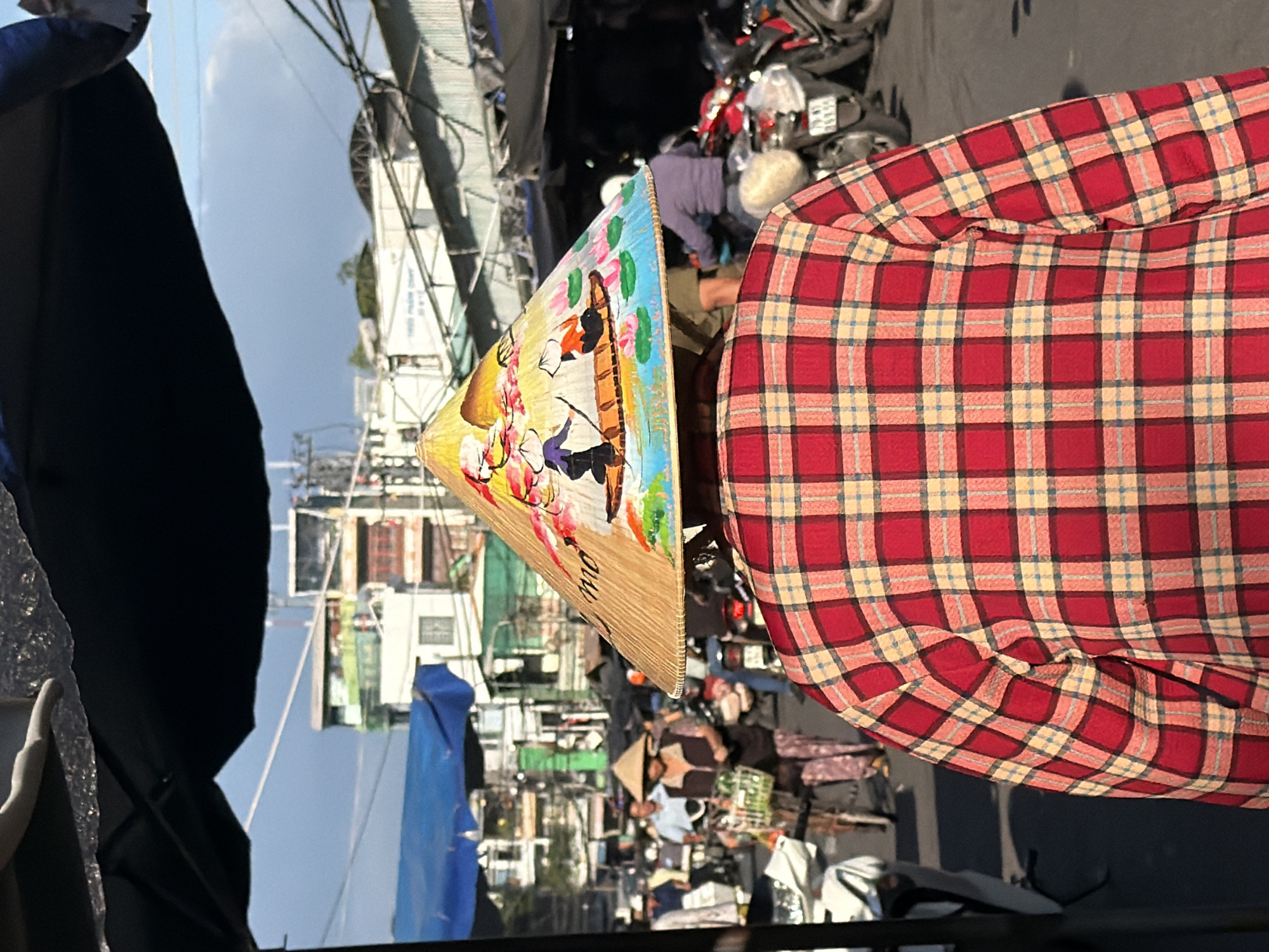 The image captures a street scene in Nha Trang, Vietnam, focusing on the back of a person wearing a red and yellow plaid shirt and a conical hat adorned with a painted scene of people in a boat on water with lotus flowers. The hat is tilted forward, obscuring the person's face. In the background, a bustling street is visible with several motorbikes, some parked and others in motion. Buildings with varying architectural styles, including some with colorful facades and balconies, line the street. A blue tarp is visible overhead, possibly part of a market stall or temporary shelter. The sky above is blue with some white clouds, indicating daylight. The overall impression is of a vibrant and active urban environment.