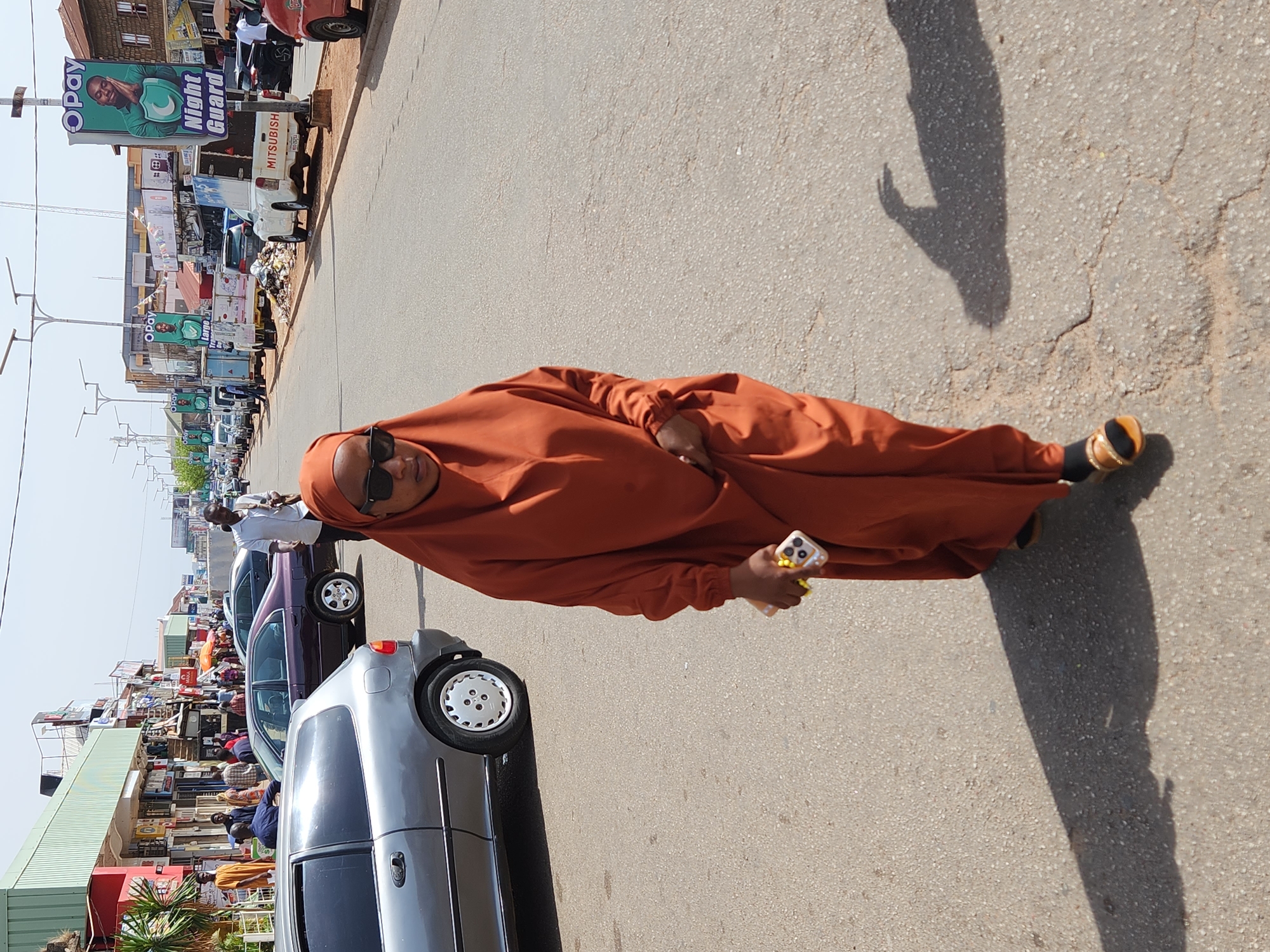 A full shot of a person on a street. The person is wearing an orange robe and sunglasses, and is holding a cell phone. They are standing in the middle of a street with parked cars on the left and buildings in the background. In the city of Unknown, Unknown. The day appears bright and sunny.