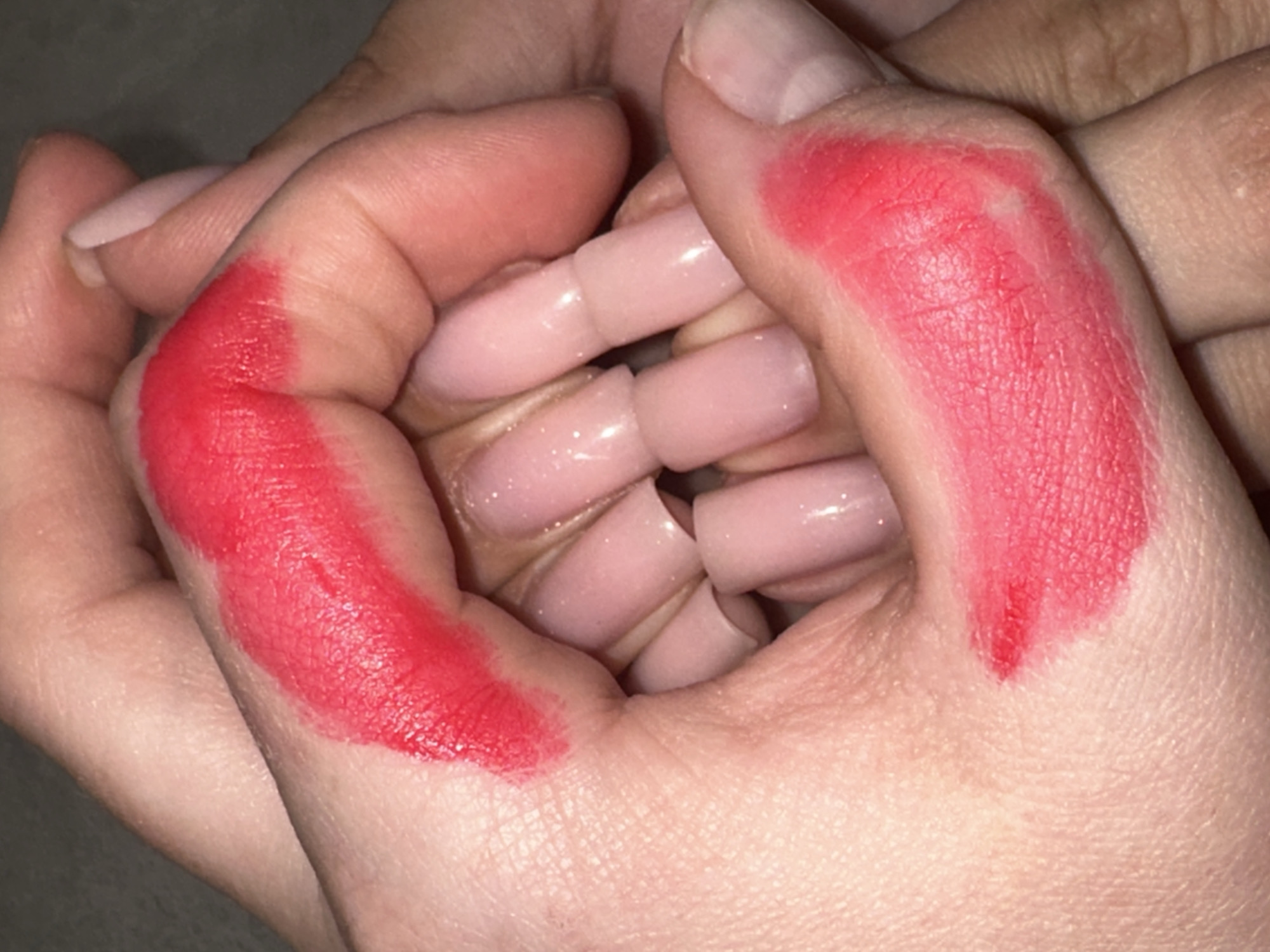 This close-up image features two intertwined hands, against a dark, out-of-focus background. One hand, positioned centrally, exhibits long, light pink or nude-colored acrylic nails with a glossy finish, suggesting artificial nail extensions. These nails appear uniform in shape, mostly square or coffin-style.
Vibrant, matte red cosmetic product, strongly resembling lipstick, is smeared across the skin of both hands. On the left side of the frame, a substantial patch of this red substance covers the knuckles and back of the hand. On the right, it is similarly applied to the knuckles and fingers. The application is visibly textured and somewhat uneven, highlighting the natural lines, creases, and pores of the fair skin beneath. In some areas, the skin's texture is clearly visible through the red substance, and a few small uncolored patches appear within the red areas.
The scene focuses exclusively on the hands and the cosmetic application, with no other subjects or objects in clear view. The activity appears to be a deliberate act of applying or swatching the red product on the hands, possibly for an artistic or promotional purpose. There are no discernible location cues within the image, nor any visible text. The time of day, weather, or emotions cannot be determined from the visual information. The provided location context is Chaikovsky, Russia, but this is not visually supported by the image itself.