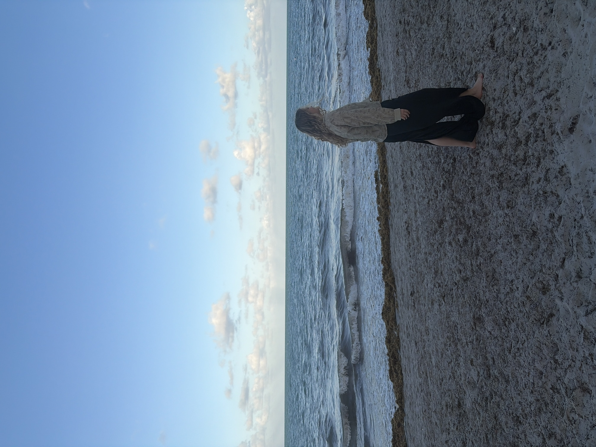 The image depicts a beach scene in Key Biscayne, United States of America. The sky is predominantly blue with scattered white clouds. The ocean is visible, with waves approaching the shoreline. A person is standing on the beach, wearing a light-colored top and dark pants. The sand appears to be wet and firm.
