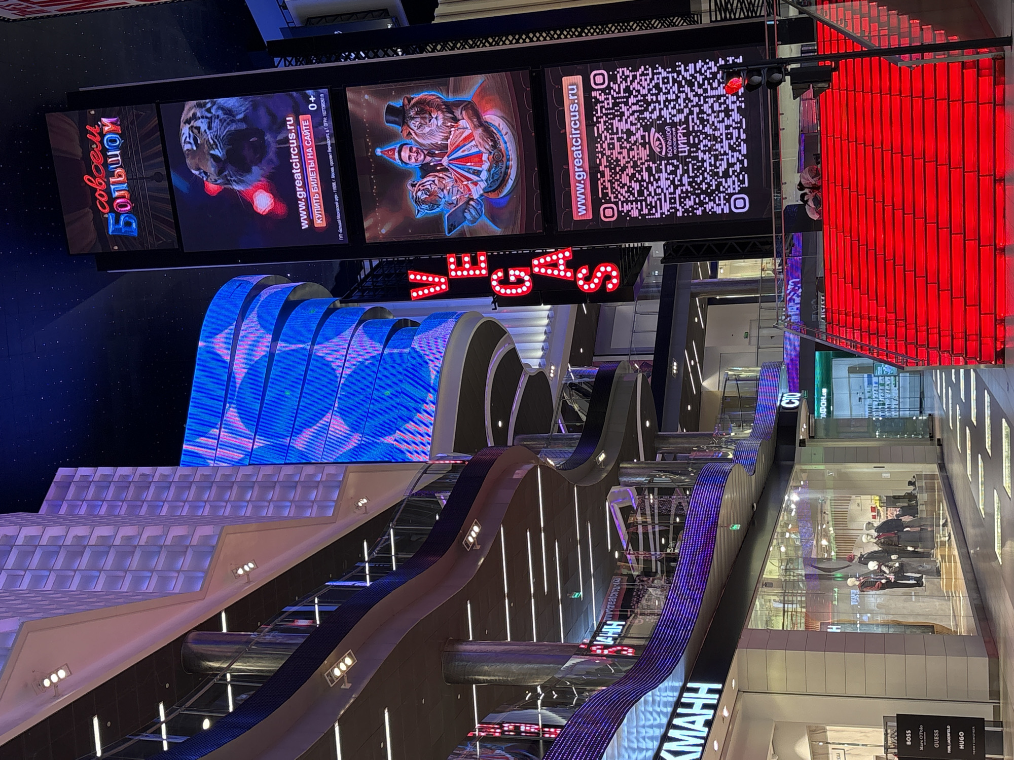 The image depicts an interior view of a modern shopping mall in Khimki, Russia. The scene is characterized by its vibrant LED lighting and large digital displays. In the foreground, a brightly lit red staircase ascends, drawing the viewer's eye upwards. On the right side of the image, a tall digital billboard displays advertisements for a circus, featuring images of a tiger, a lion tamer, and a QR code. The text on the billboard is in Russian and advertises "www.greatcircus.ru" and "Купить билеты на сайте" (Buy tickets on the website). Above this, a neon sign spells out "VEGAS".
Further into the mall, curved architectural elements with blue and purple LED lighting are visible, suggesting a futuristic or entertainment-themed design. Several storefronts are visible, including one with mannequins displayed in a window. The overall ambiance is one of a lively and modern commercial space, possibly a large entertainment complex or mall. There are a few people visible, some sitting on the red staircase and others walking in the background. The ceiling appears to be a dark expanse dotted with lights, mimicking a night sky. The image captures a dynamic and visually stimulating environment within the mall.